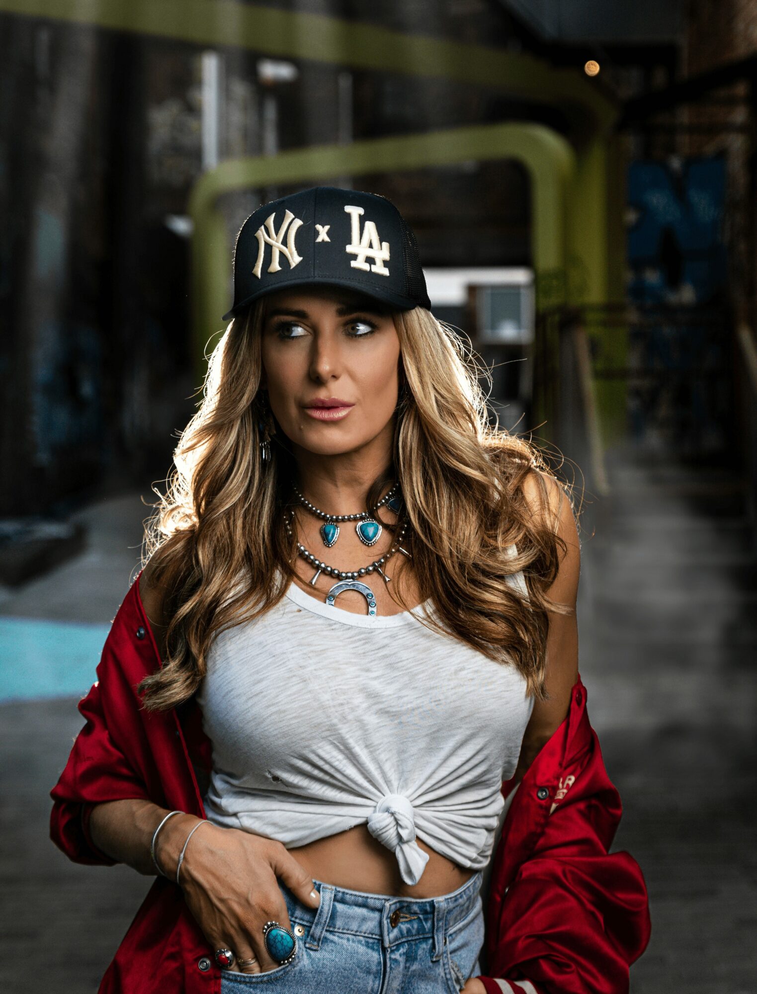 Woman with long wavy hair wearing a black cap, white knotted top, red jacket, and jewelry, standing outdoors.