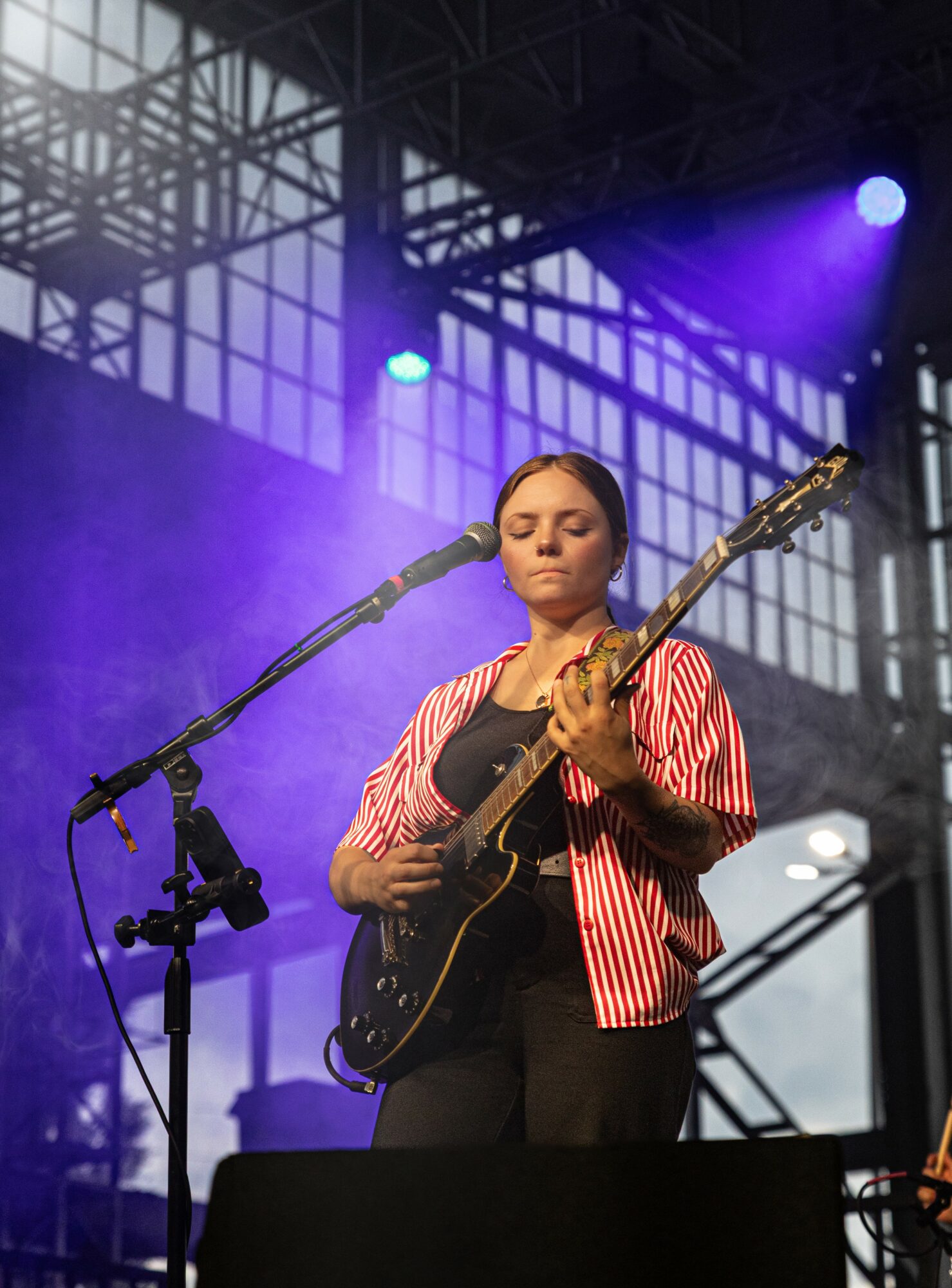 A woman plays an electric guitar on stage with purple lighting and a metal framework background.