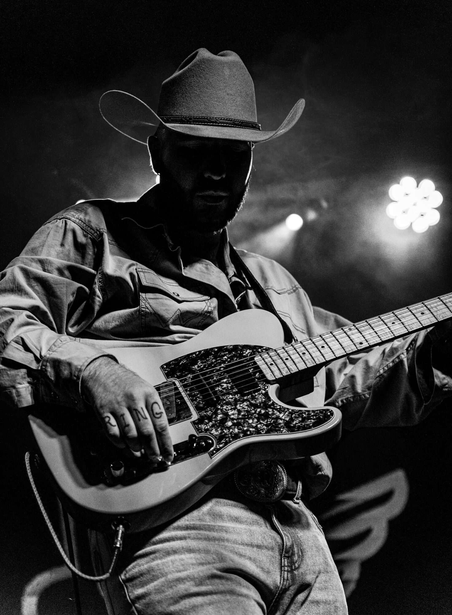 Man wearing cowboy hat playing electric guitar on stage with bright lights behind him.