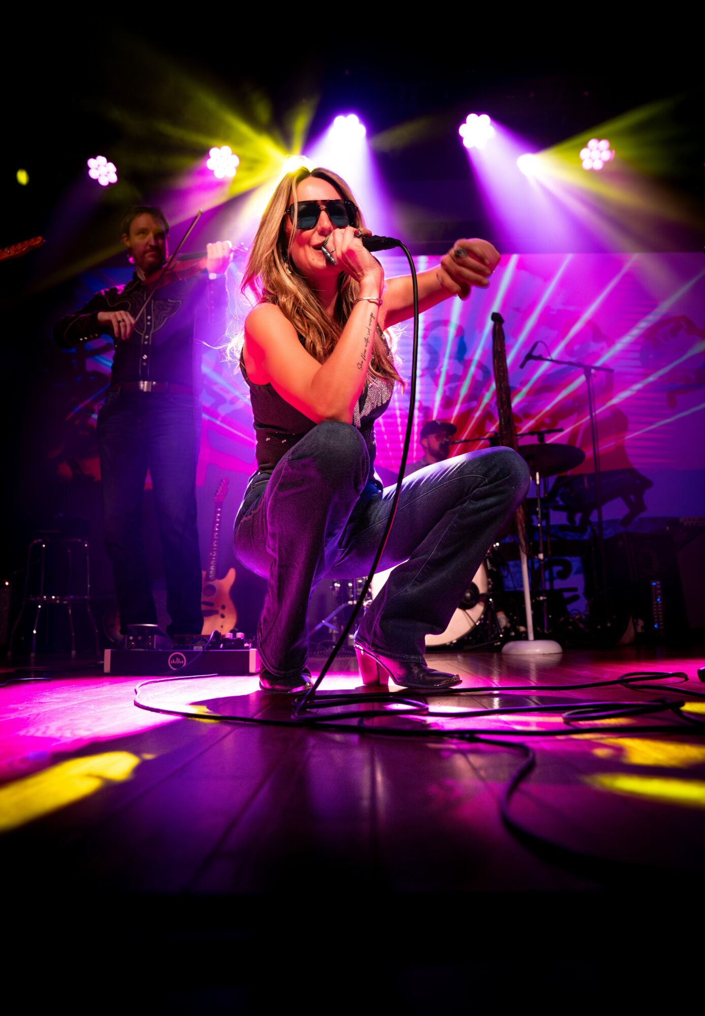 Female singer crouching with microphone on stage, colorful lights shining behind her, band members in background.