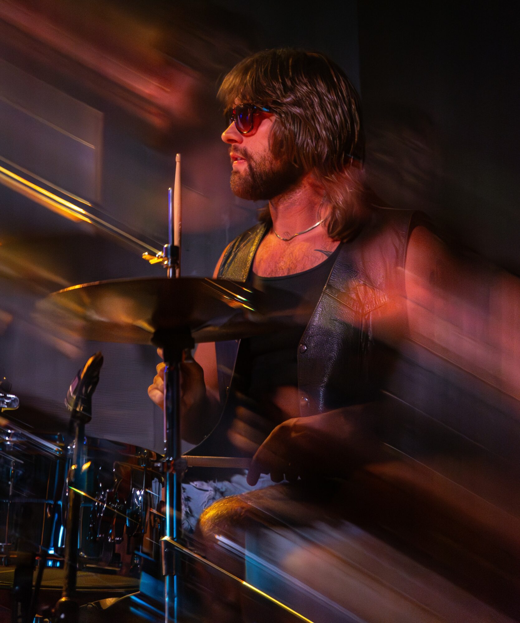 Musician playing drums with motion blur, wearing sunglasses and a sleeveless shirt, in a dark setting.