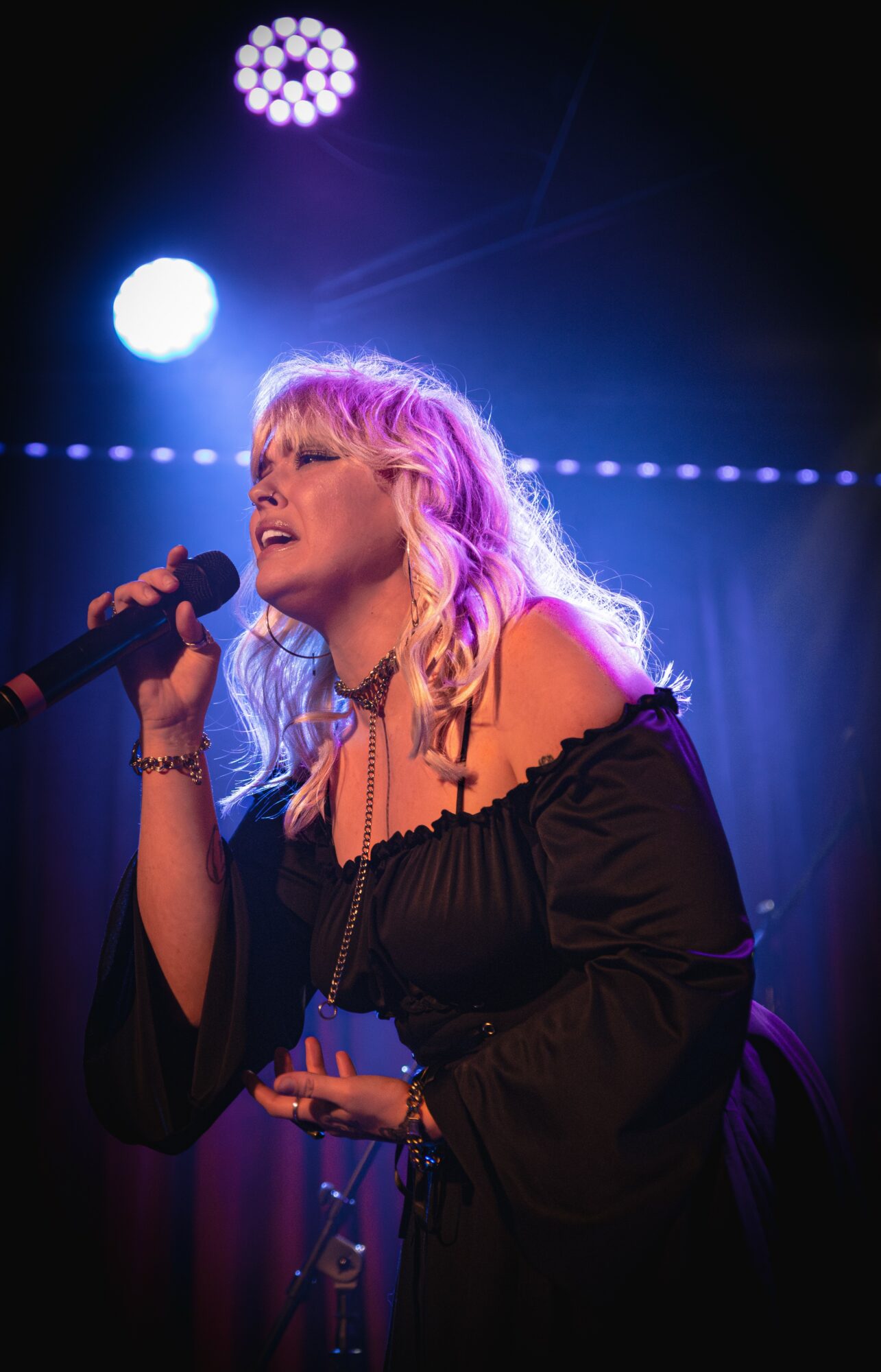 Female singer with blonde hair performs on stage under colorful lights, holding a microphone, wearing a black off-shoulder dress.