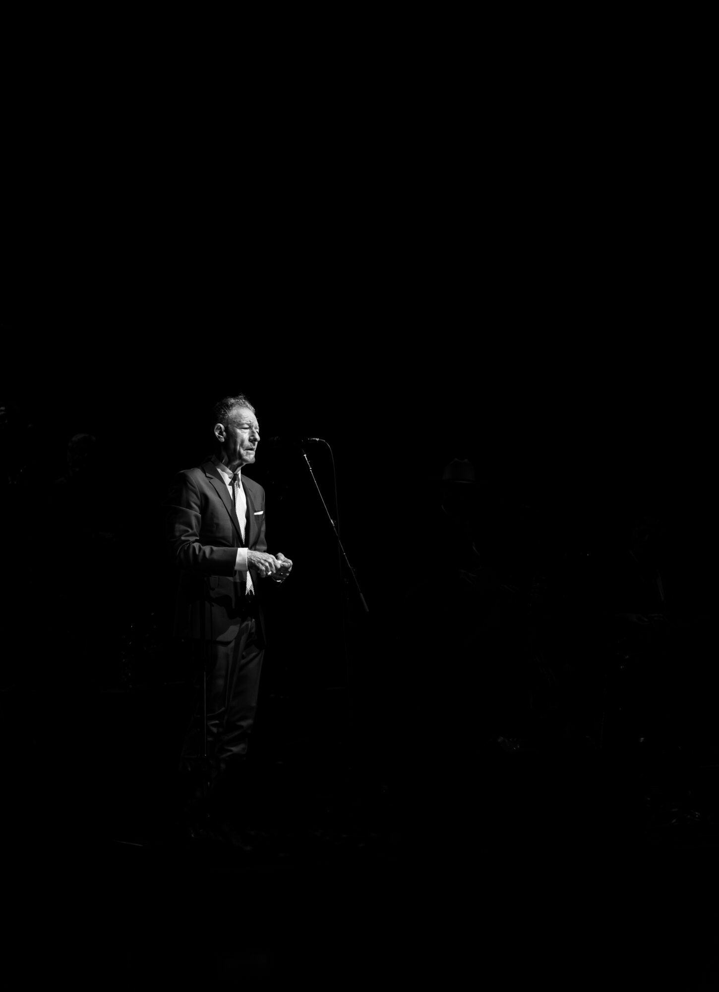 Person standing on stage with microphone, illuminated by a spotlight, in a dark background.