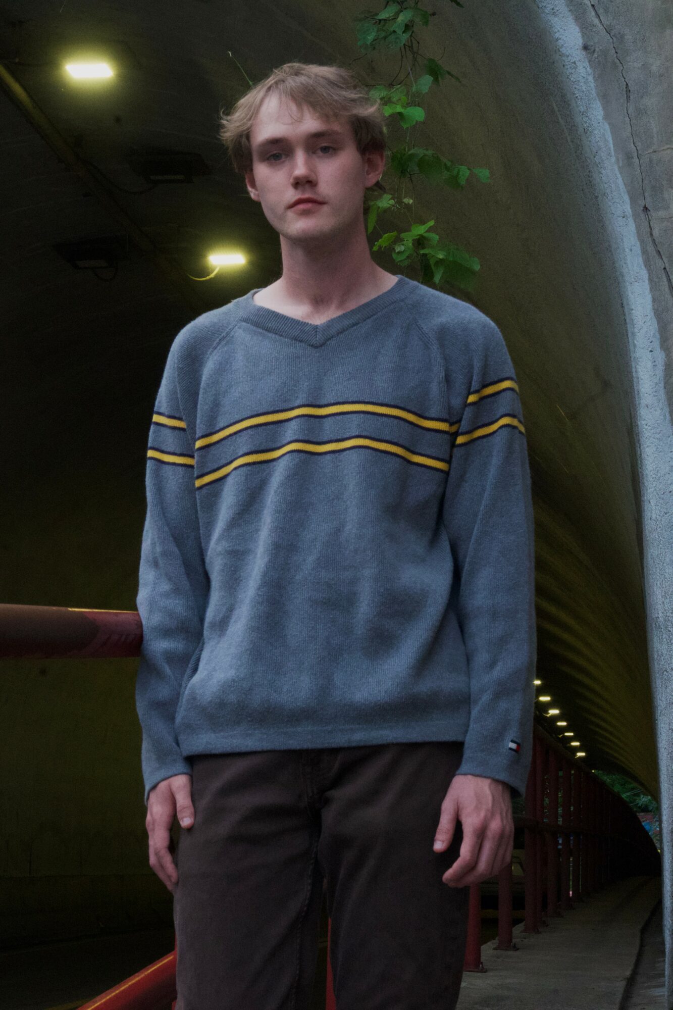 Young person with curly hair wearing a blue sweater with yellow stripes, standing outdoors near a tunnel entrance.