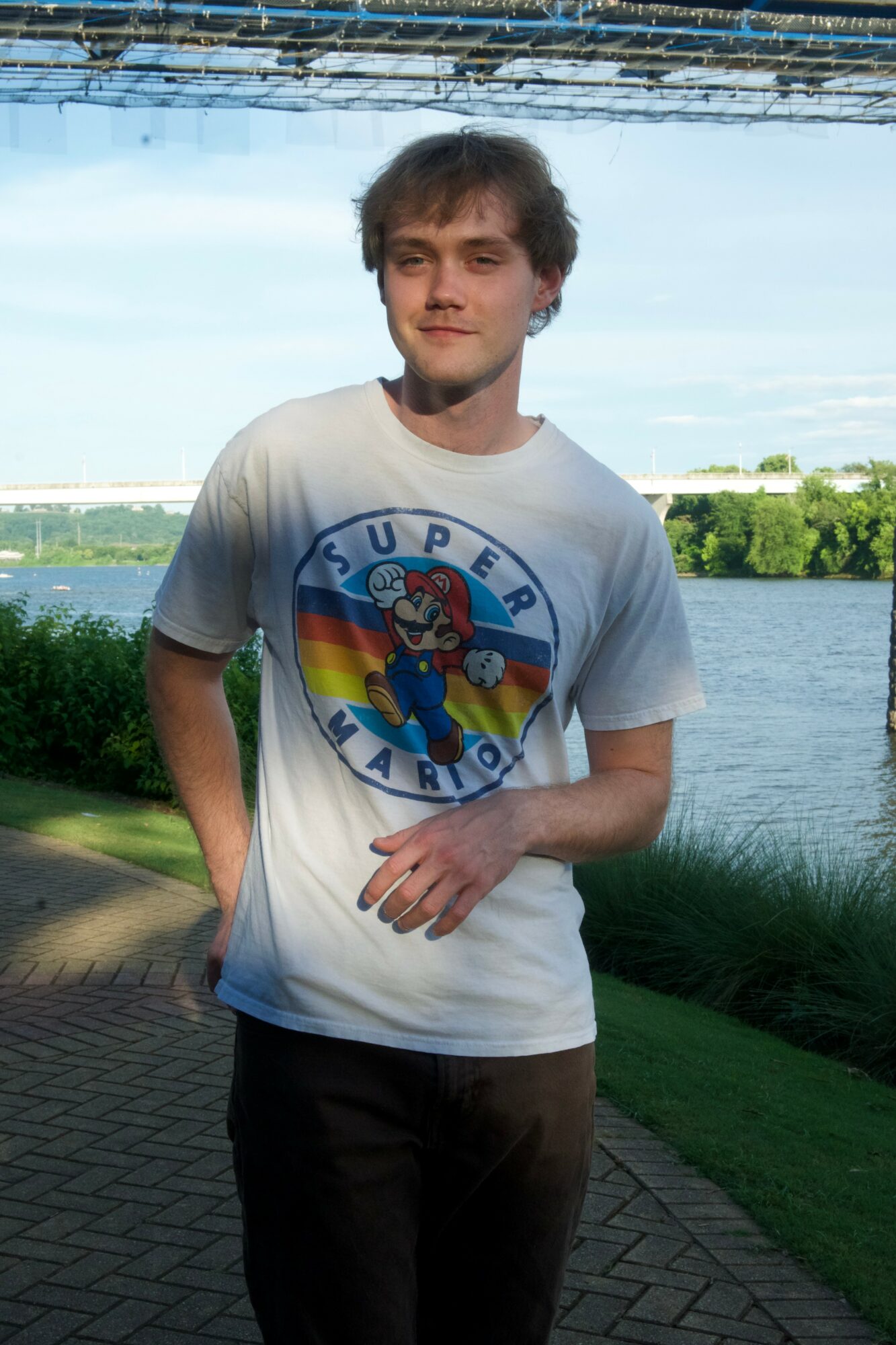 Young man standing outdoors near water with trees and sky in background, wearing a Super Mario T-shirt, smiling.