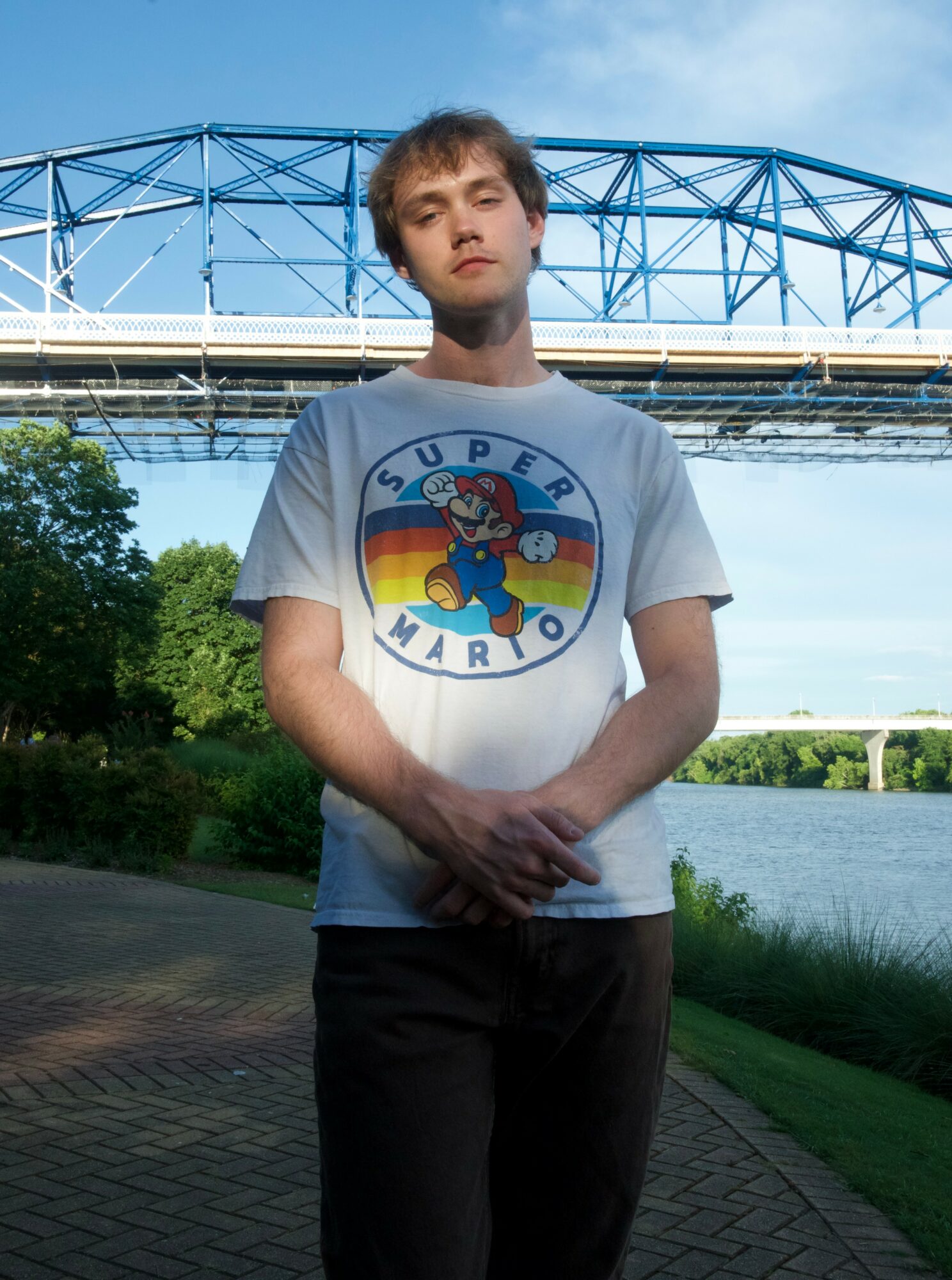 Young man standing outdoors near water with a bridge in the background, wearing a Super Mario T-shirt, hands clasped in front.