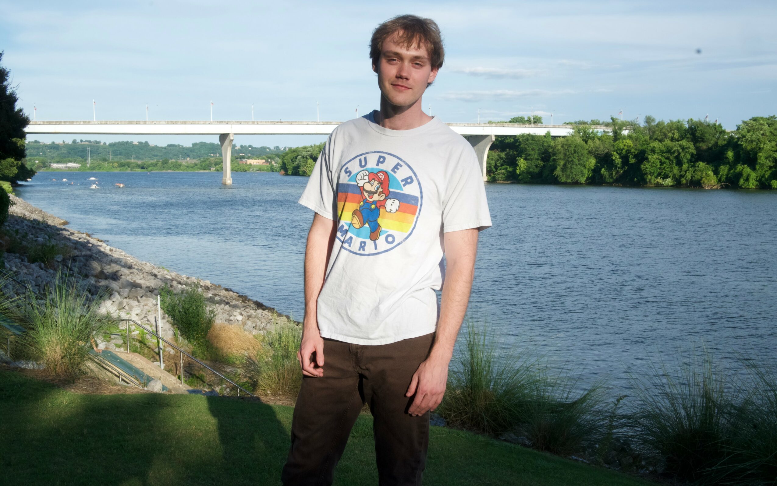Young man standing outdoors near a river with a bridge in the background, wearing a white T-shirt and dark pants.
