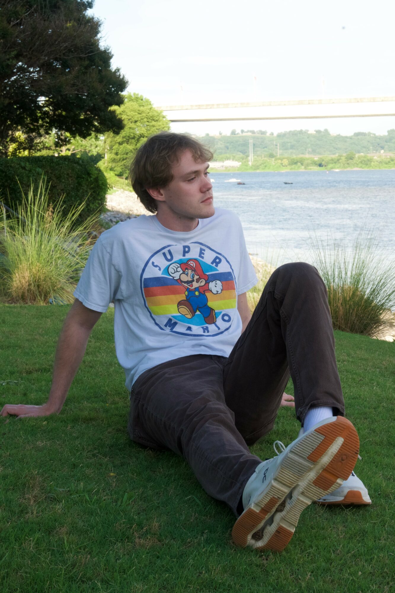 Person sitting on grass near water with trees and sky in background, wearing a colorful T-shirt and dark pants.