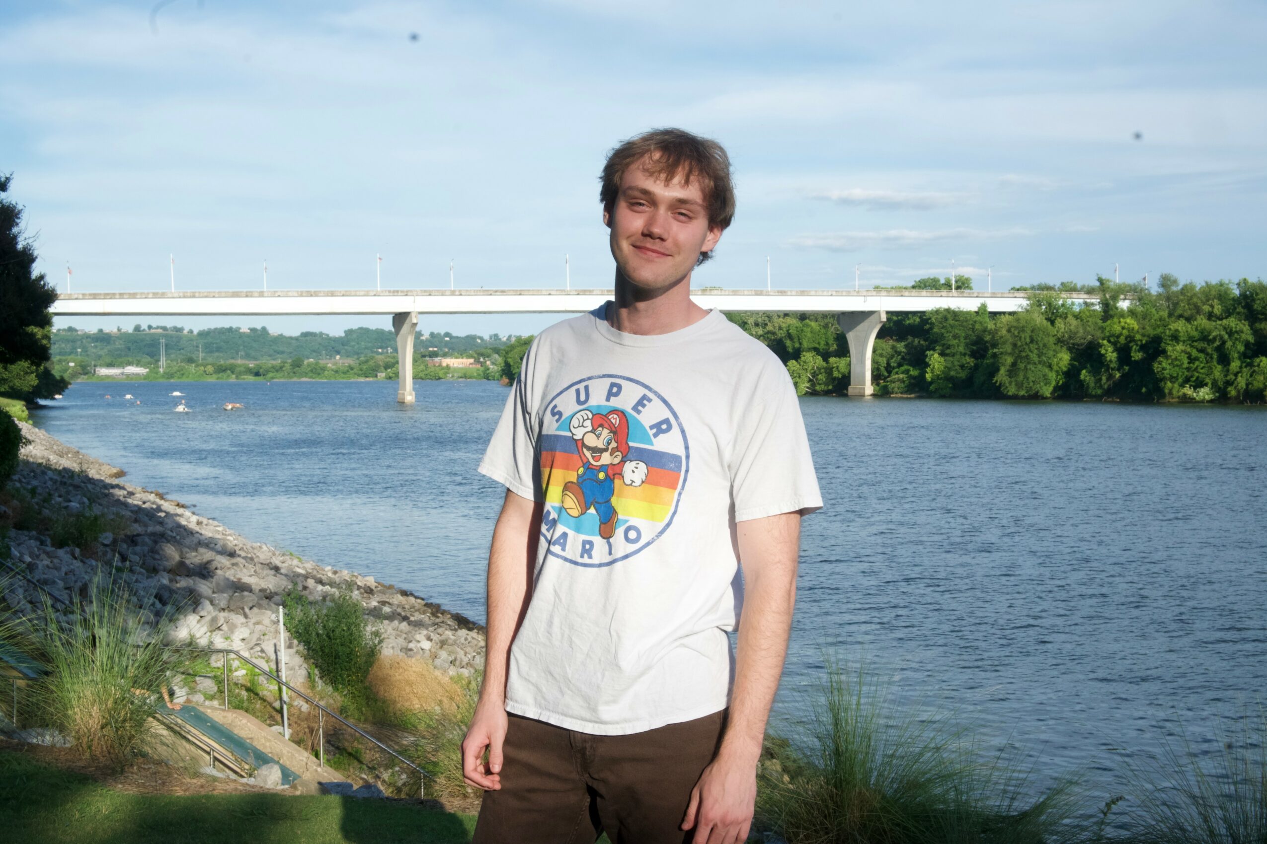 Young man standing outdoors near a river with a bridge in the background, wearing a white T-shirt and brown pants.