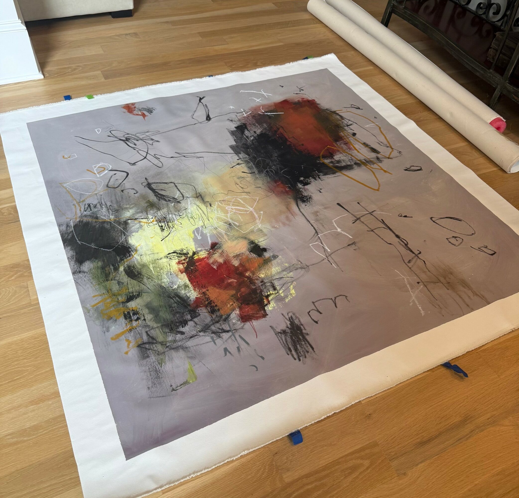 Abstract painting with black, red, yellow, and gray colors on paper, placed on a wooden floor with some rolled paper nearby.