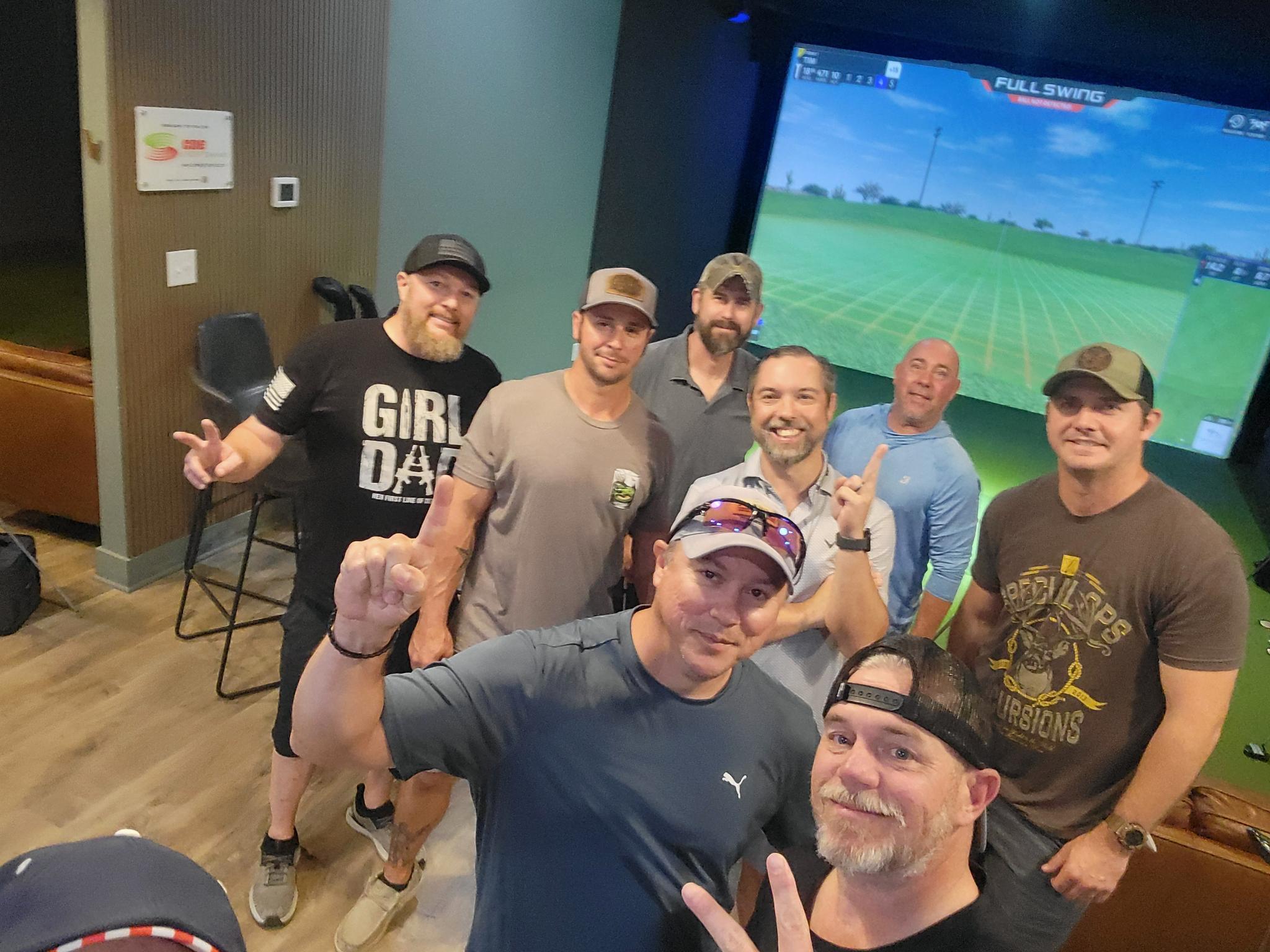 Group of seven men smiling and posing indoors with a large screen showing a golf game in the background.