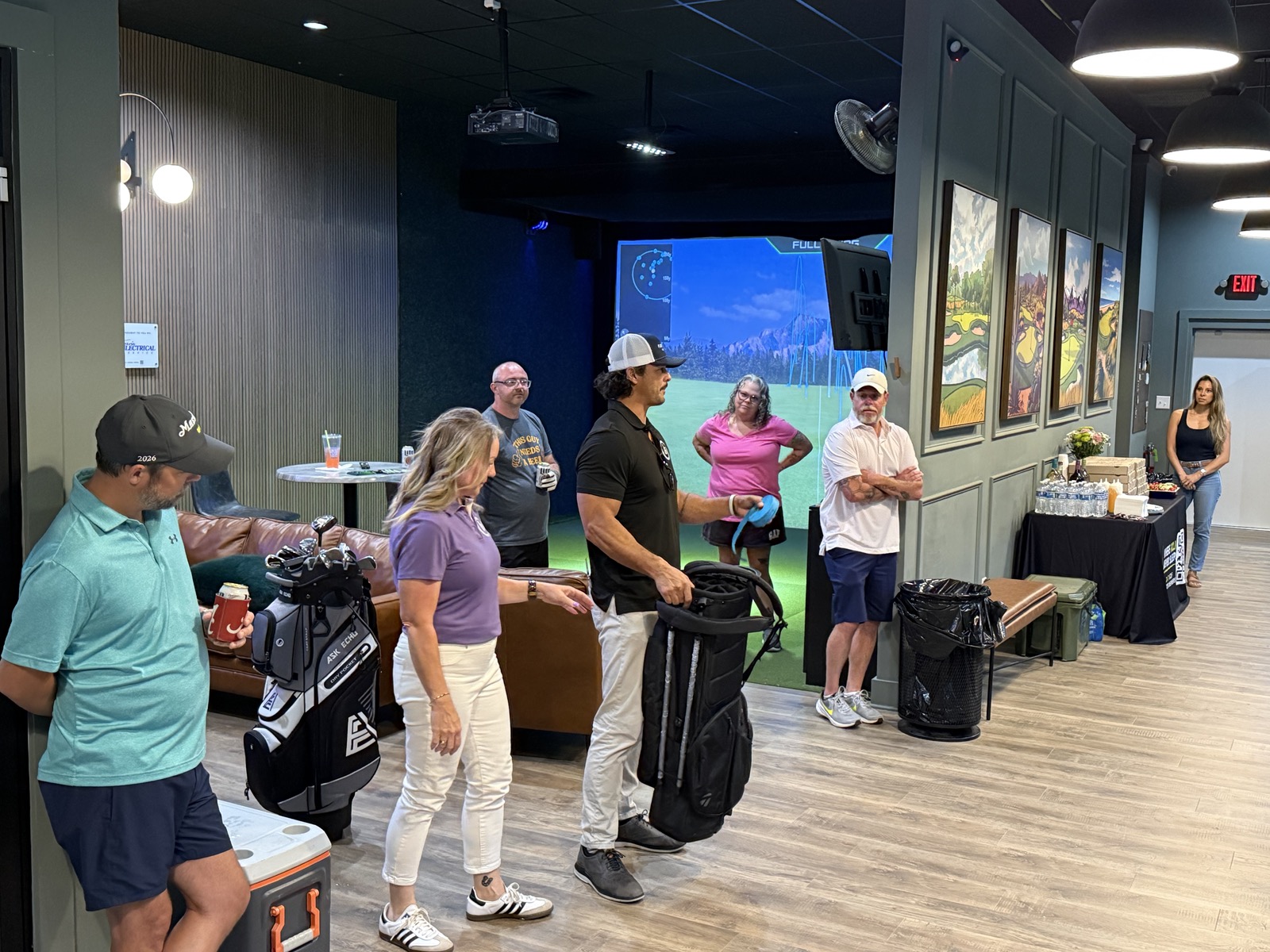 Group of people standing in a room with a large screen showing a landscape, some holding luggage, in a casual setting.