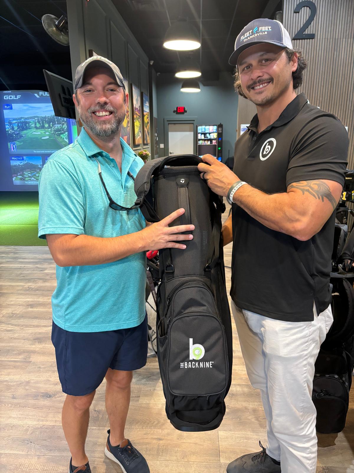Two men standing indoors, smiling, holding a golf bag between them, with screens and a sign in the background.