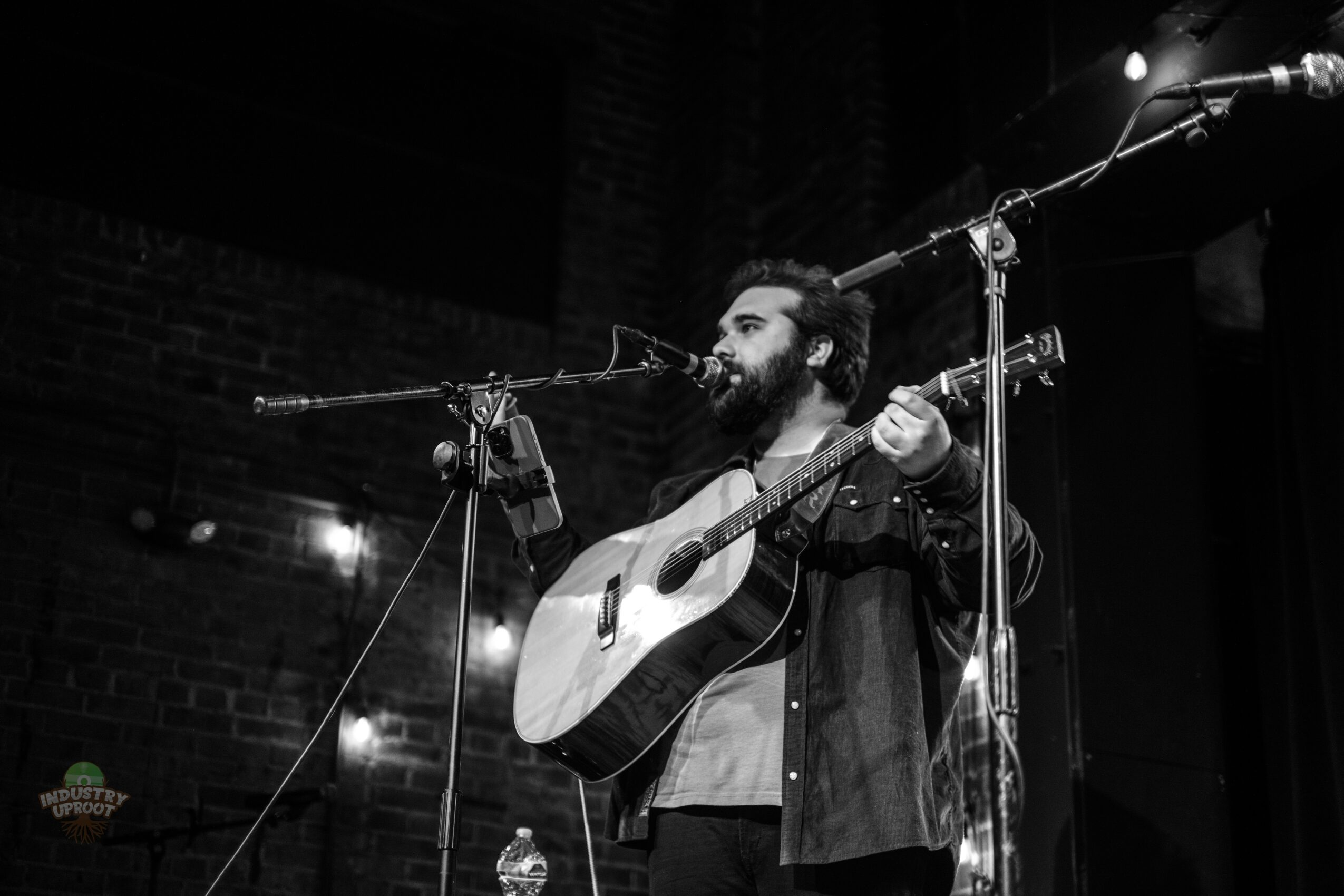 A bearded man with curly hair playing an acoustic guitar on stage with microphones, in black and white.