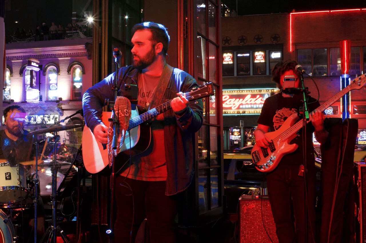 Three musicians perform on stage at night with city lights and neon signs in the background.