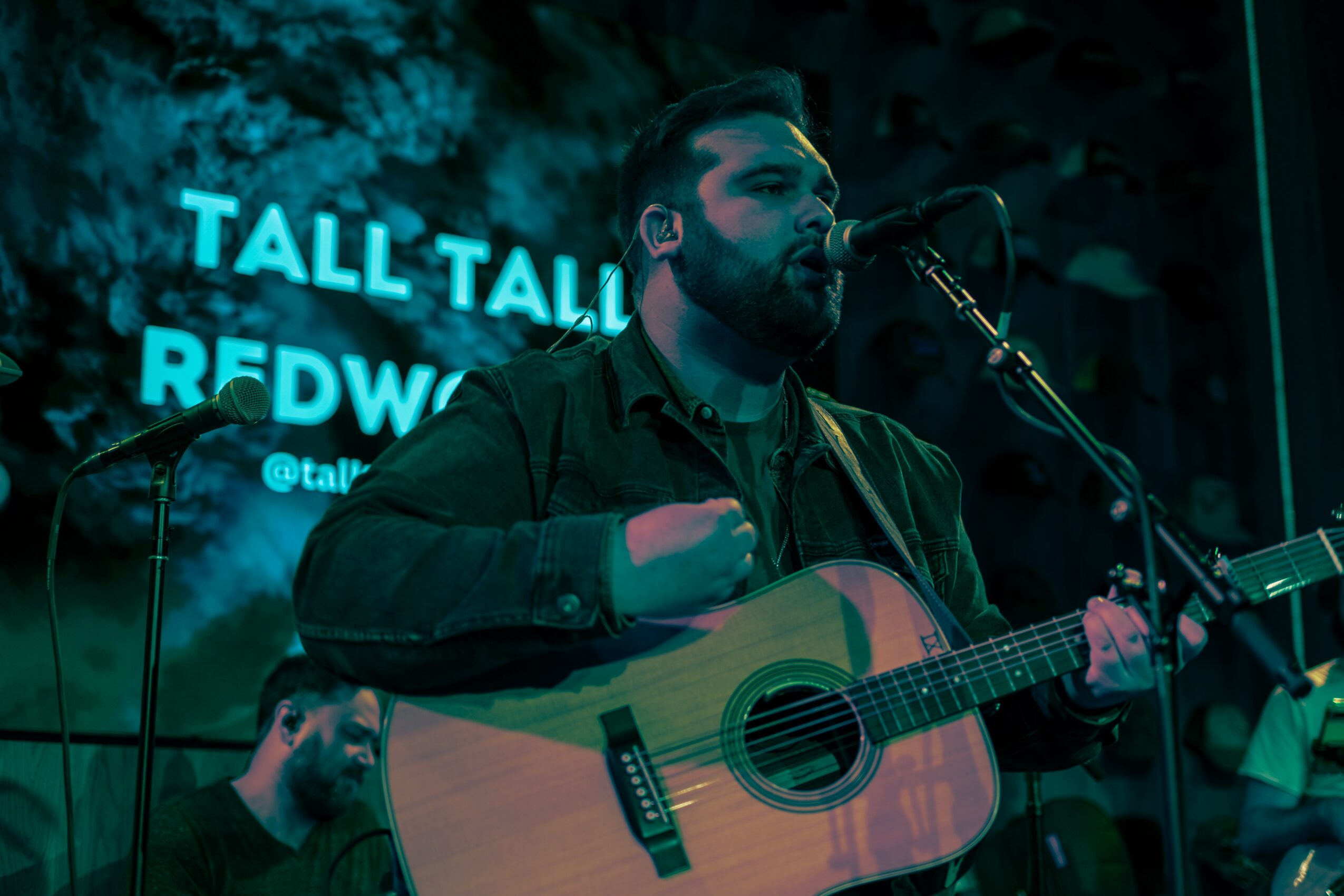 Man playing guitar and singing into microphone on stage with blue neon sign in background