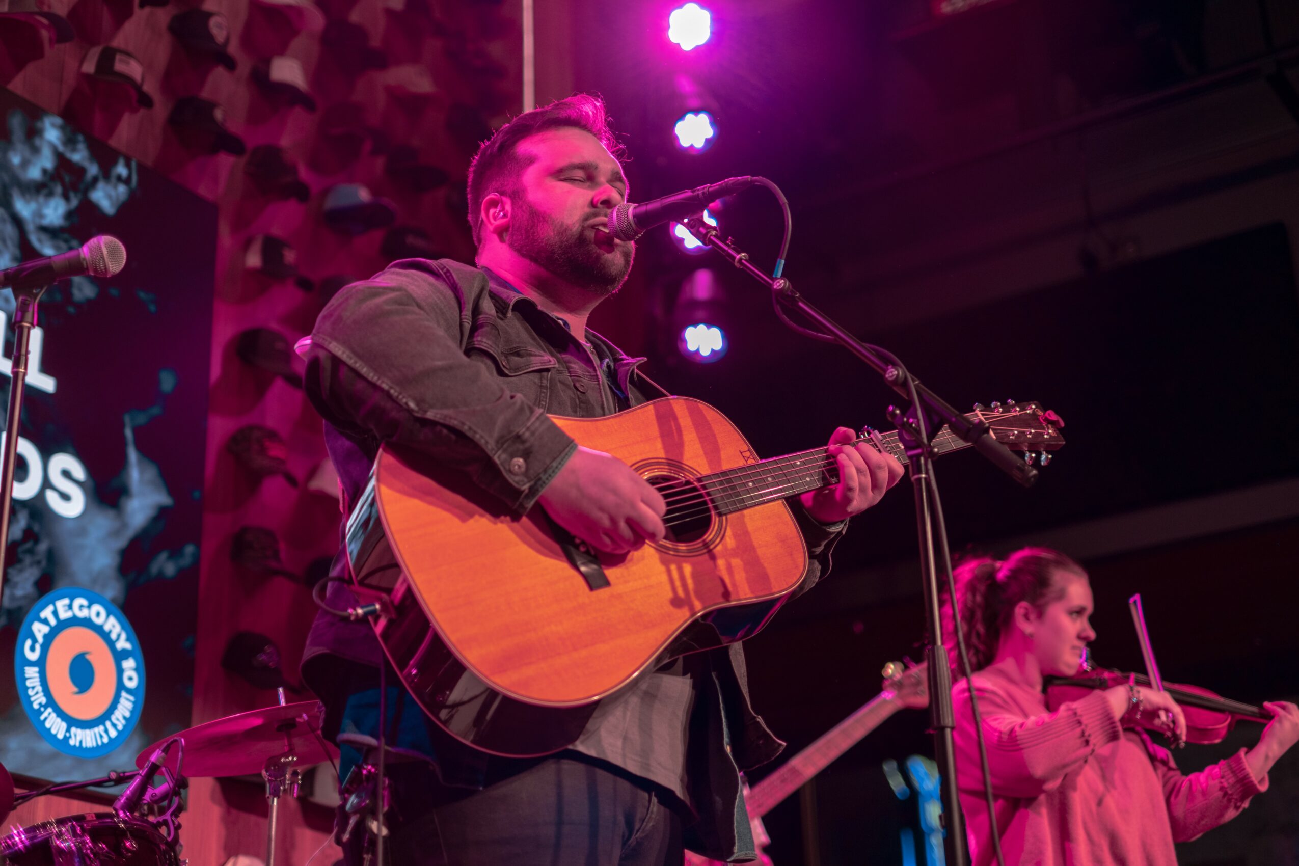 Man playing acoustic guitar and singing on stage with purple lighting, a woman playing violin in the background.