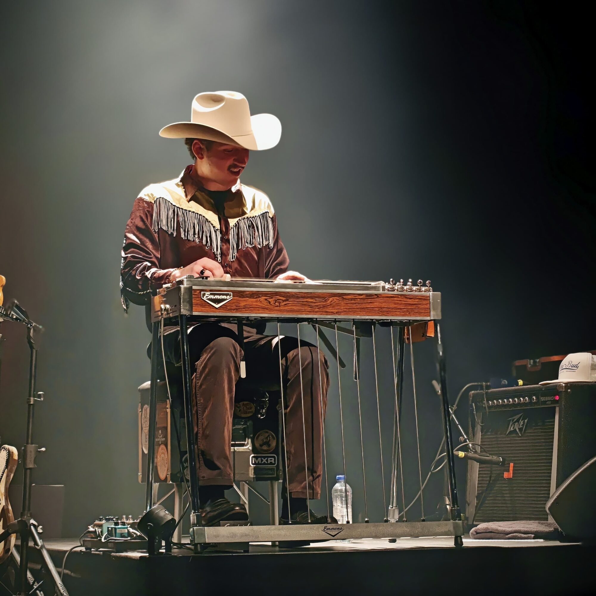 Person wearing a cowboy hat playing a pedal steel guitar on stage with equipment around them.
