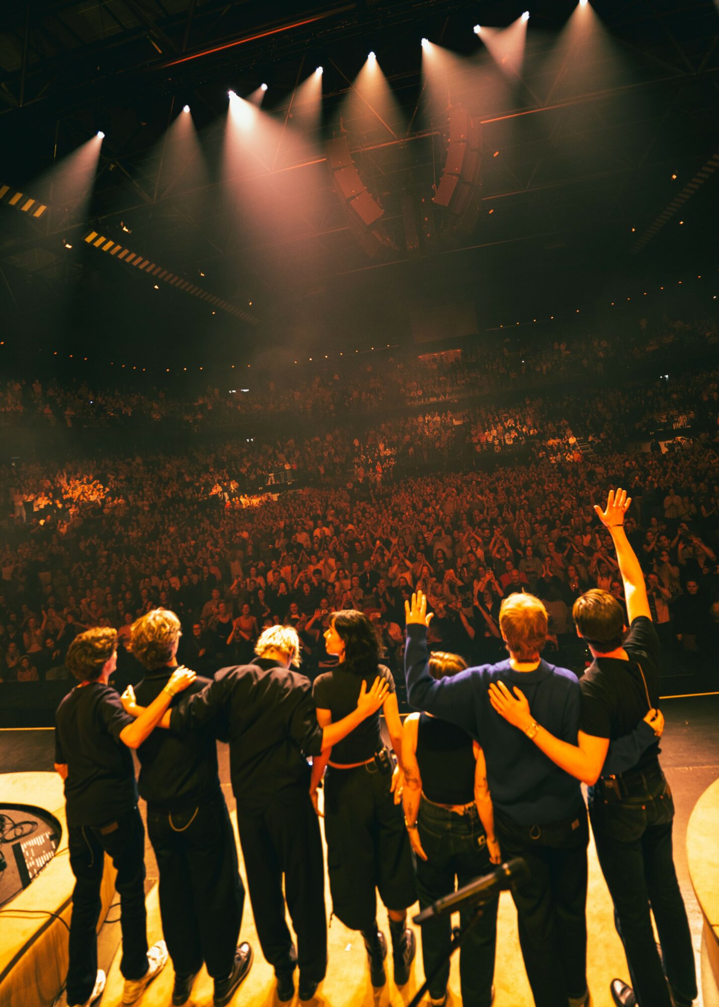 Group of people on stage facing a large audience, some with hands raised, in a theater or concert hall.