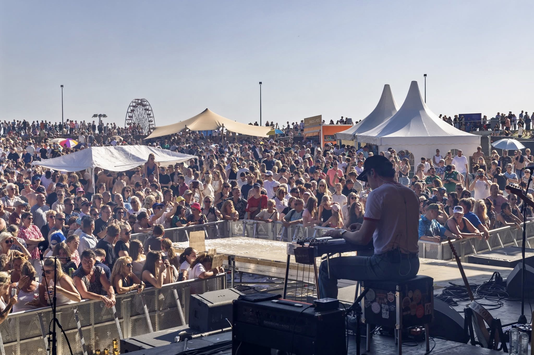 Crowd gathered at outdoor event with tents and a person performing on stage, under a clear sky.