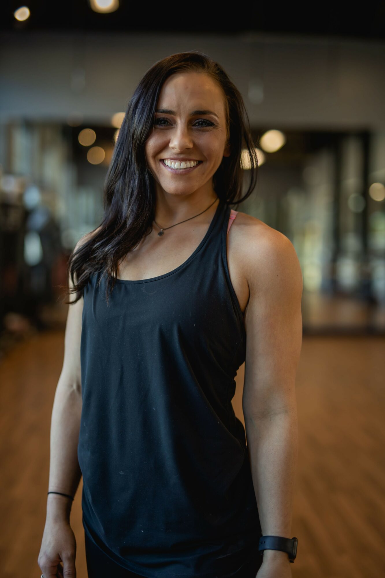 Smiling woman with long dark hair in a black tank top in a gym, wooden floor, and blurred background.