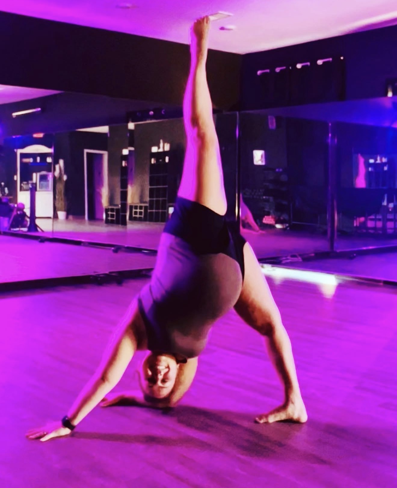 Person performing a headstand with one leg extended upward in a dance studio, purple lighting, wooden floor, mirrors, and seating area.