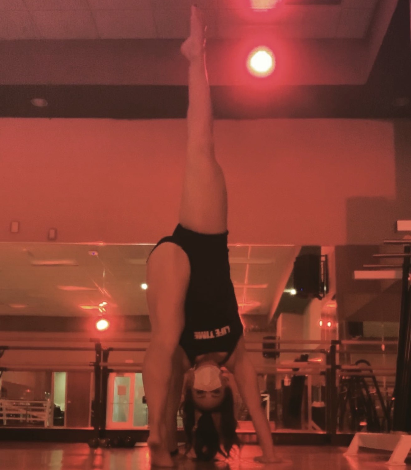 Person performing a handstand in a gym, legs extended upward, head near the floor, with gym equipment in background.
