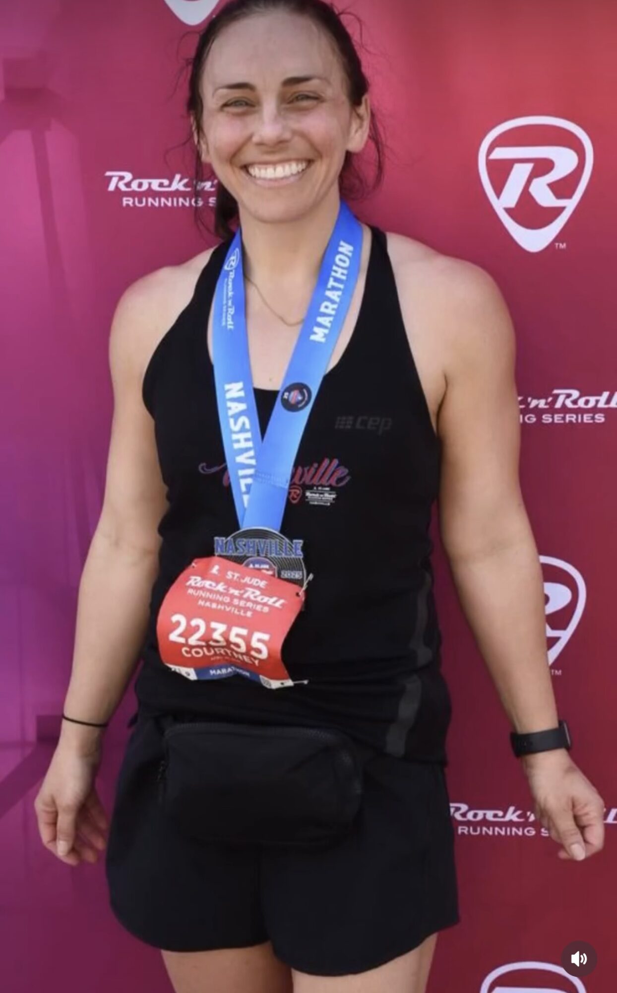 Smiling woman with a medal and race bib, standing in front of a pink background with logos.