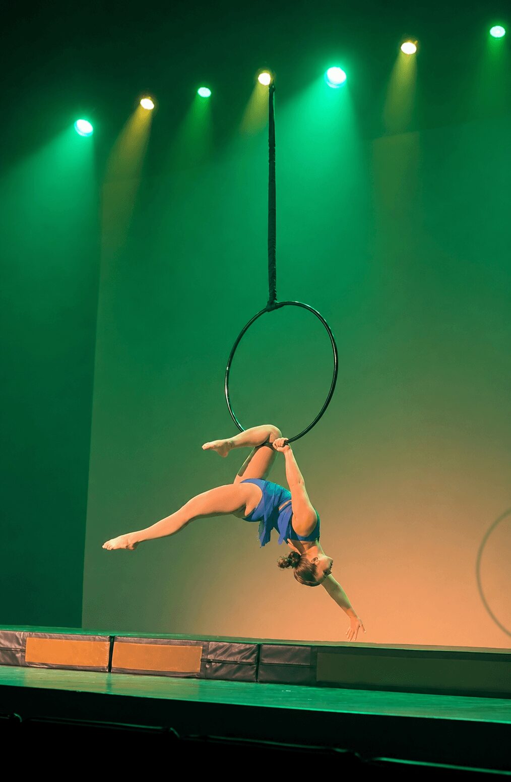 Aerial performer hanging upside down from a hoop, with stage lights and green background, performing a dance move.