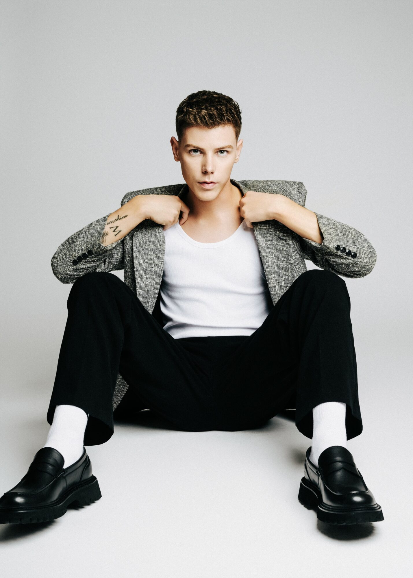Young person sitting with legs apart, wearing a gray blazer, white shirt, black pants, and black shoes, against a plain background.