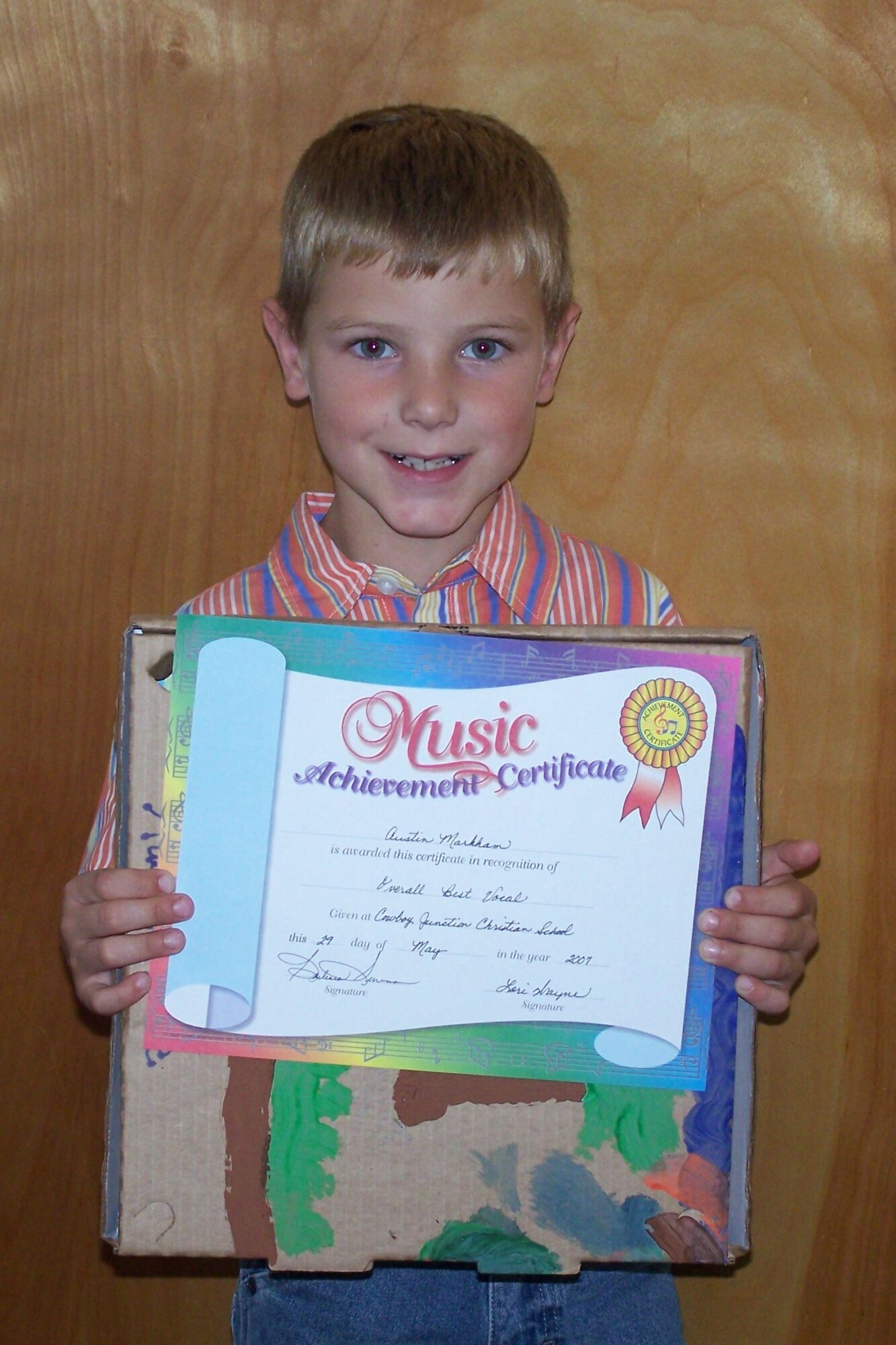 Young boy holding a colorful certificate and a box, smiling, standing in front of a wooden background.