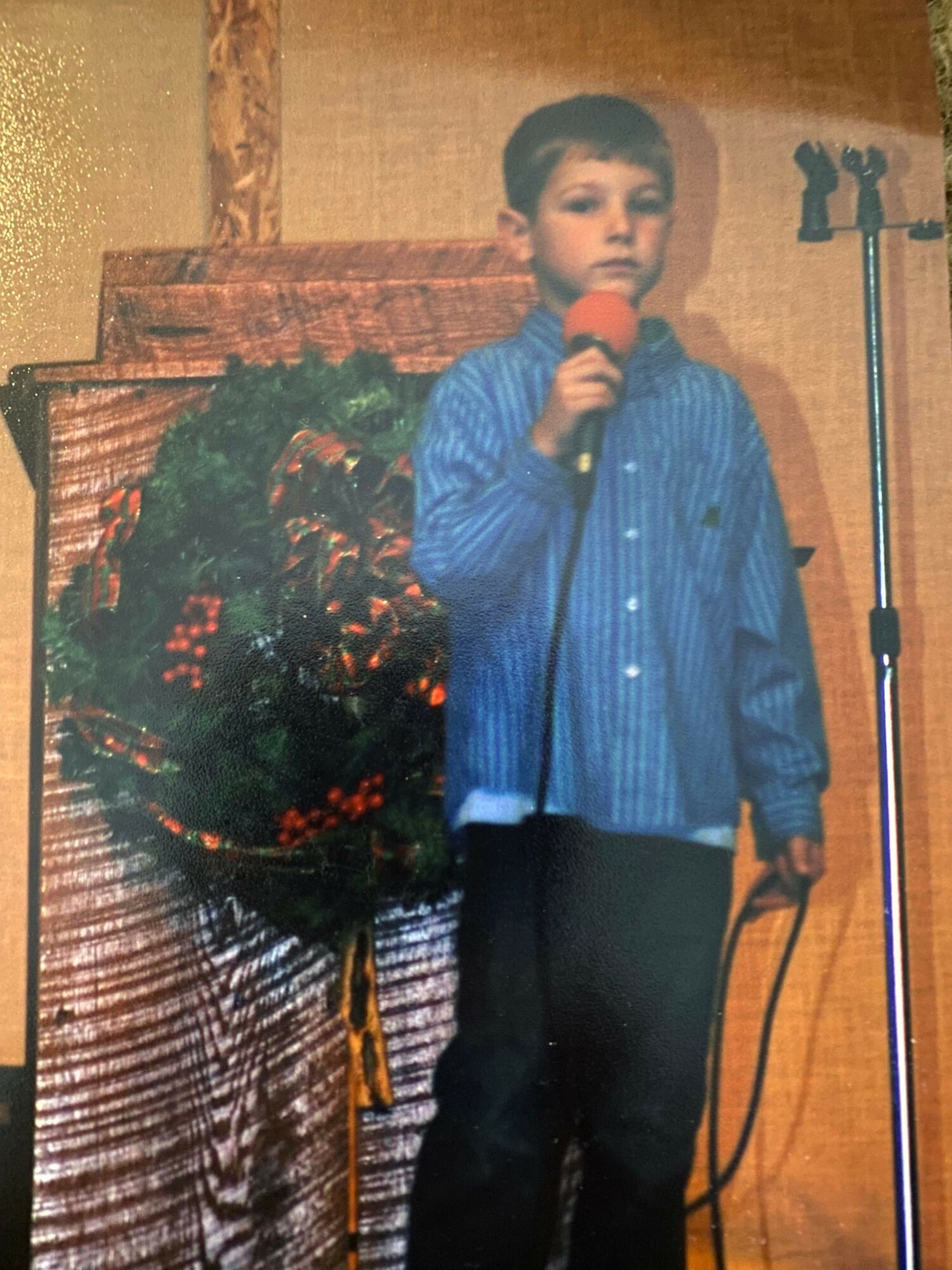 Person standing on stage holding a microphone, wearing a blue shirt and black pants, with a decorated background.