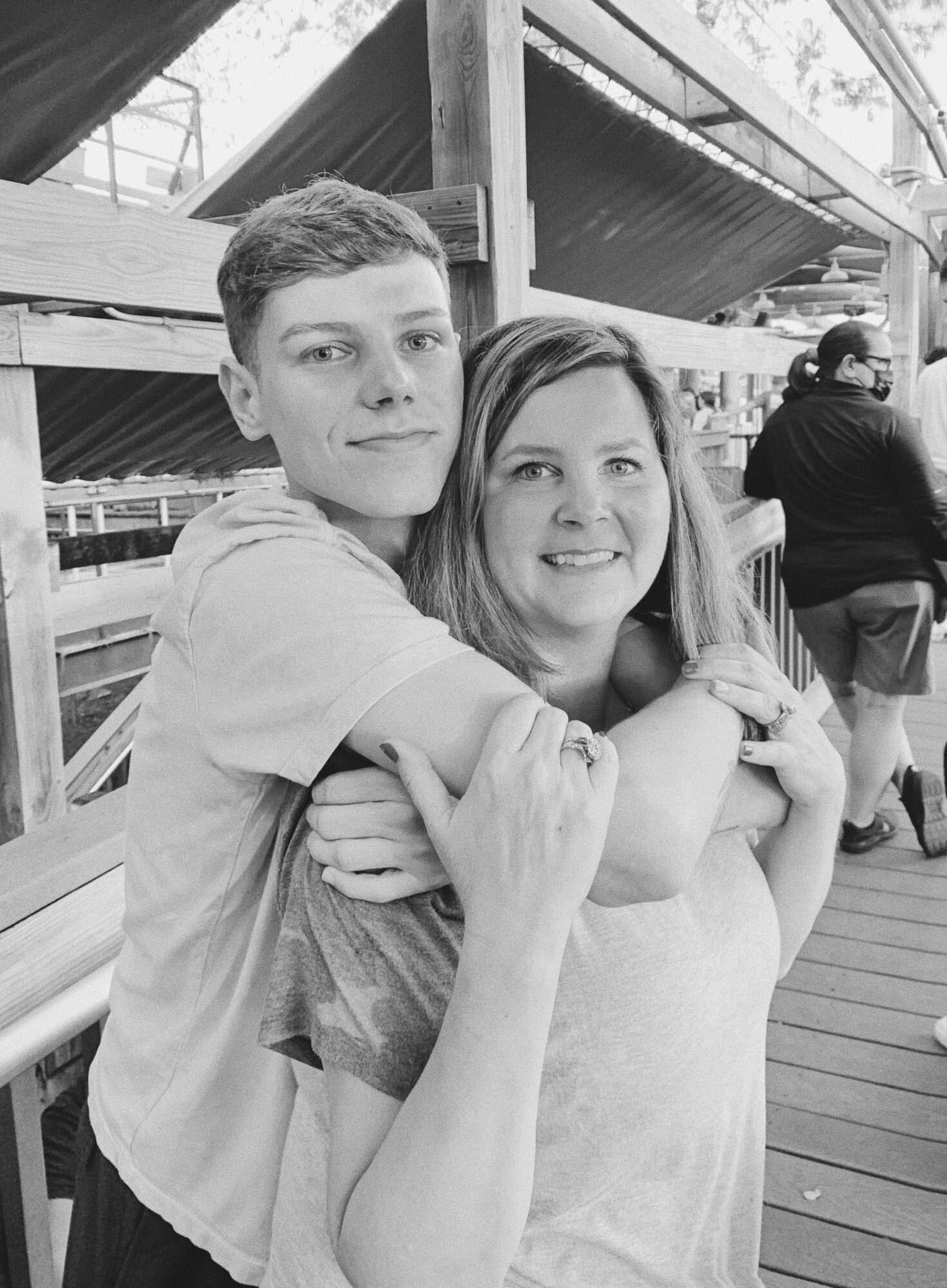 A young man and woman are hugging and smiling on a wooden deck, with boats and people in the background.