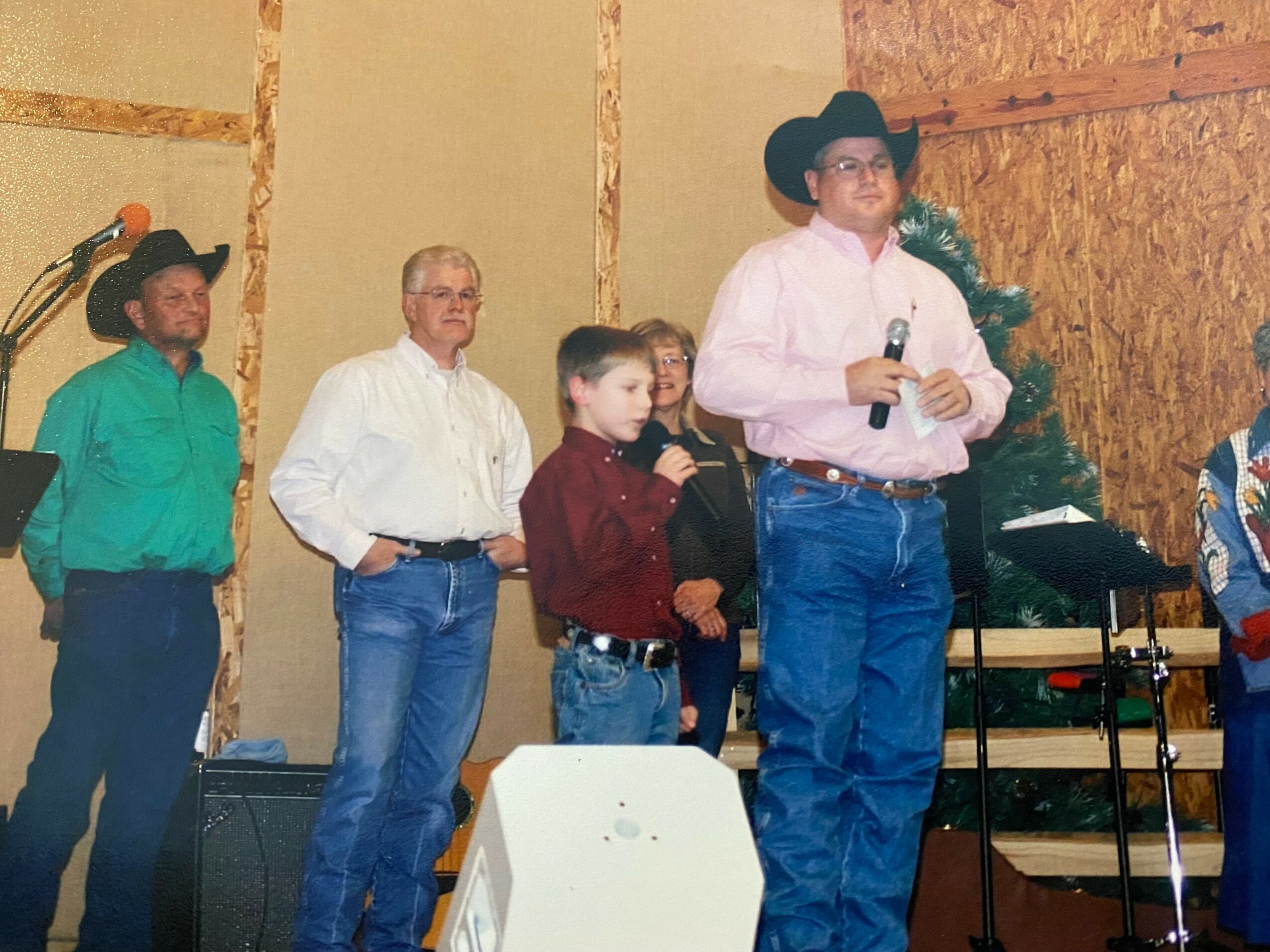Group of people standing on stage, one person holding a microphone, in a rustic setting with a Christmas tree.