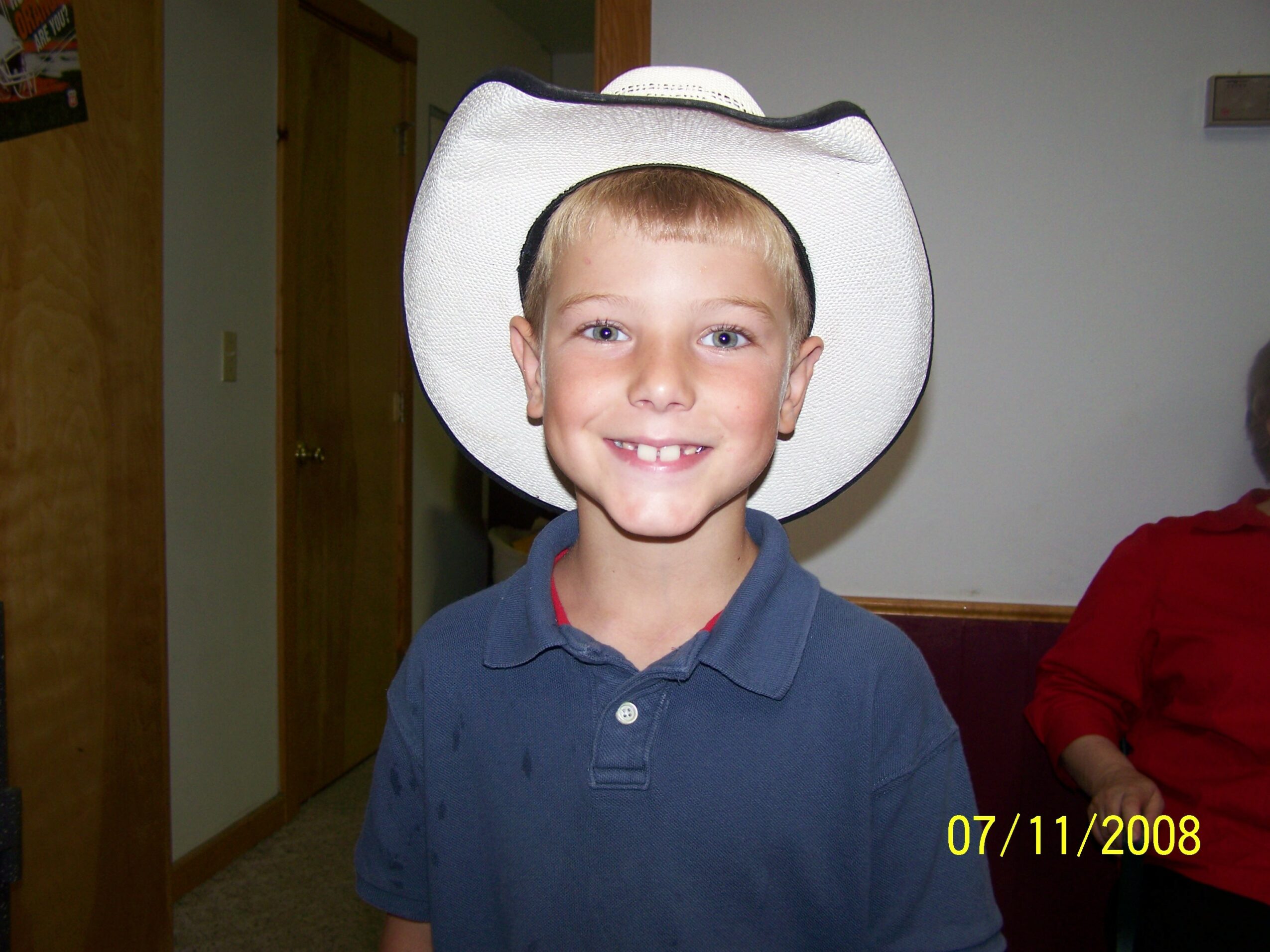 Young boy wearing a white cowboy hat and blue shirt, smiling, indoors.