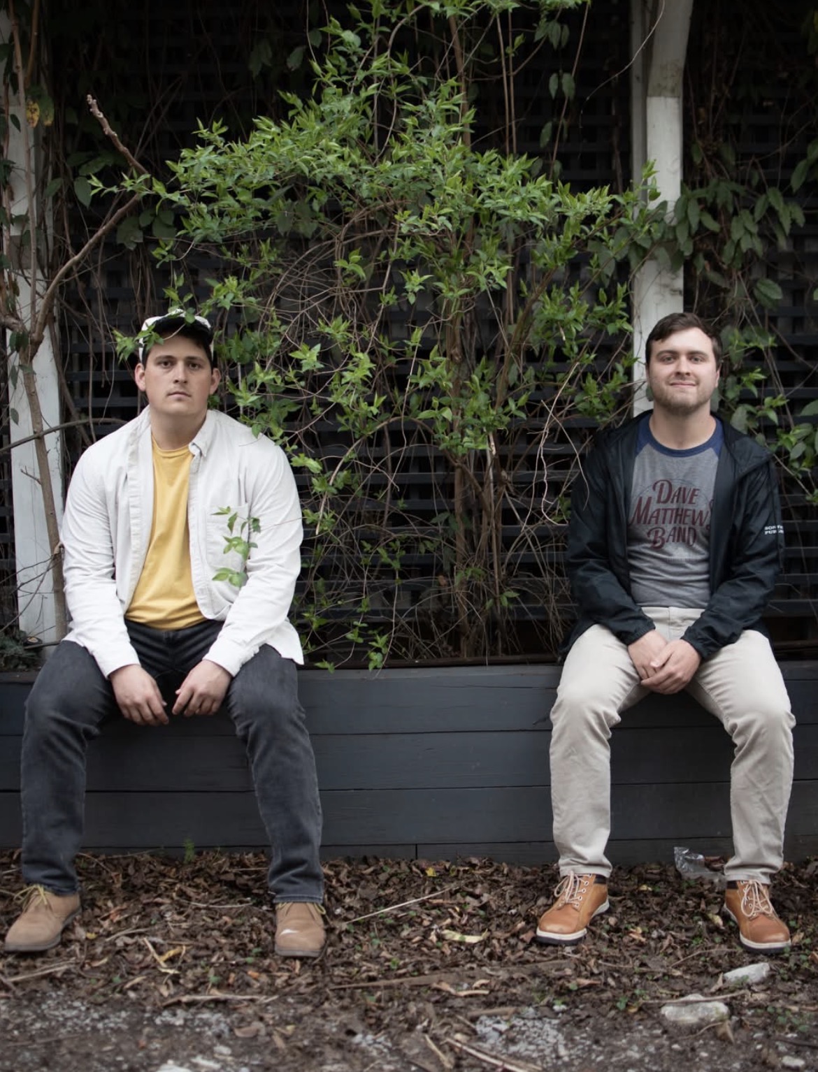 Two young men sit on a bench outdoors with greenery and a wooden fence behind them.