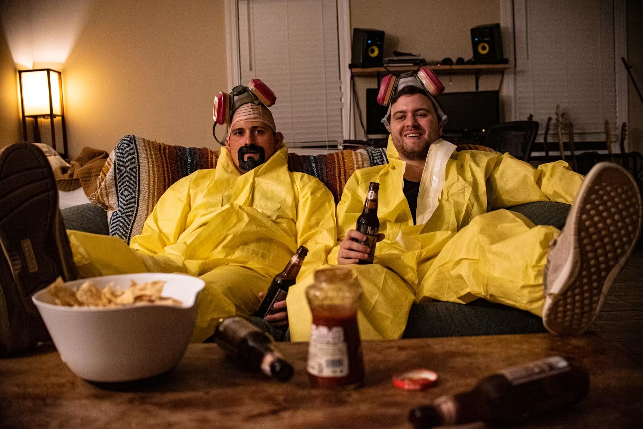 Two people in yellow hazmat suits and hair curlers sitting on a couch, holding bottles, with snacks and condiments on a table.
