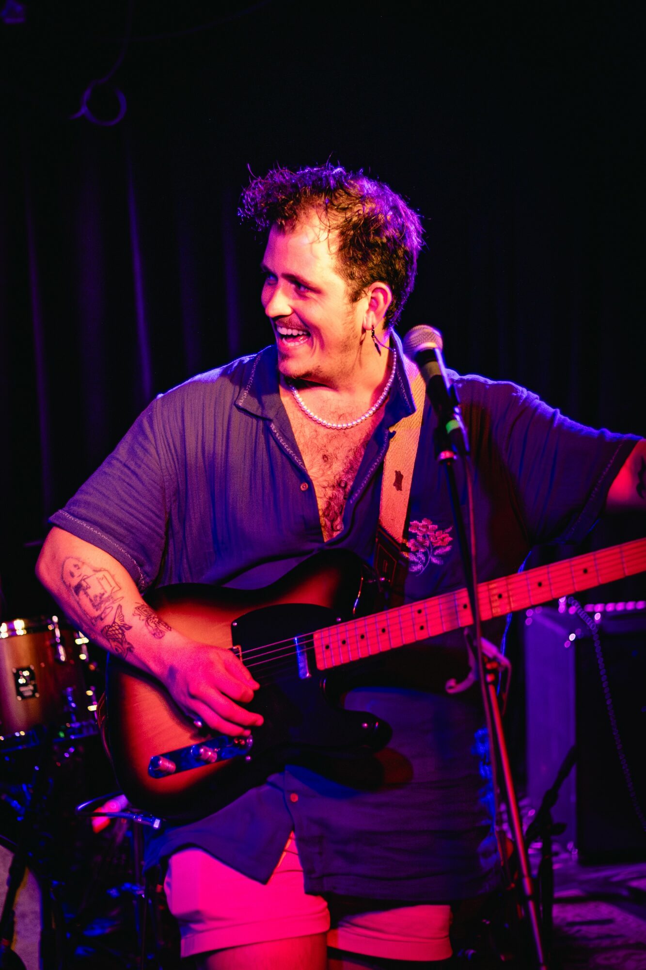 Person playing guitar on stage, smiling, with dark background and stage lighting.