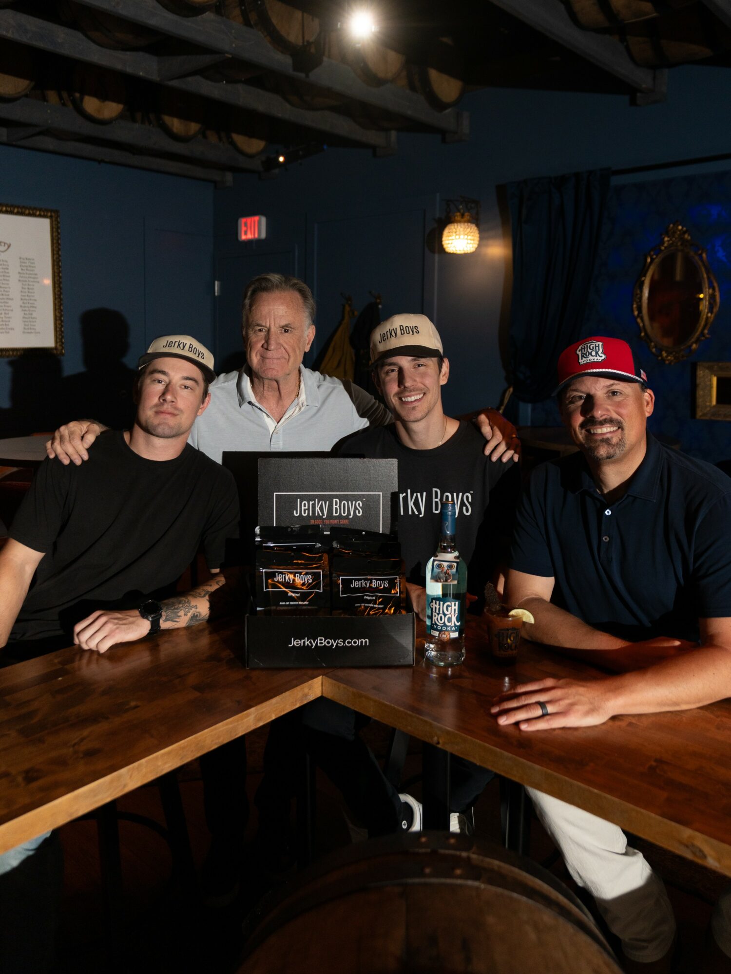 Four men sit at a bar, with a display of jerky products, in a dimly lit room with blue walls and curtains.