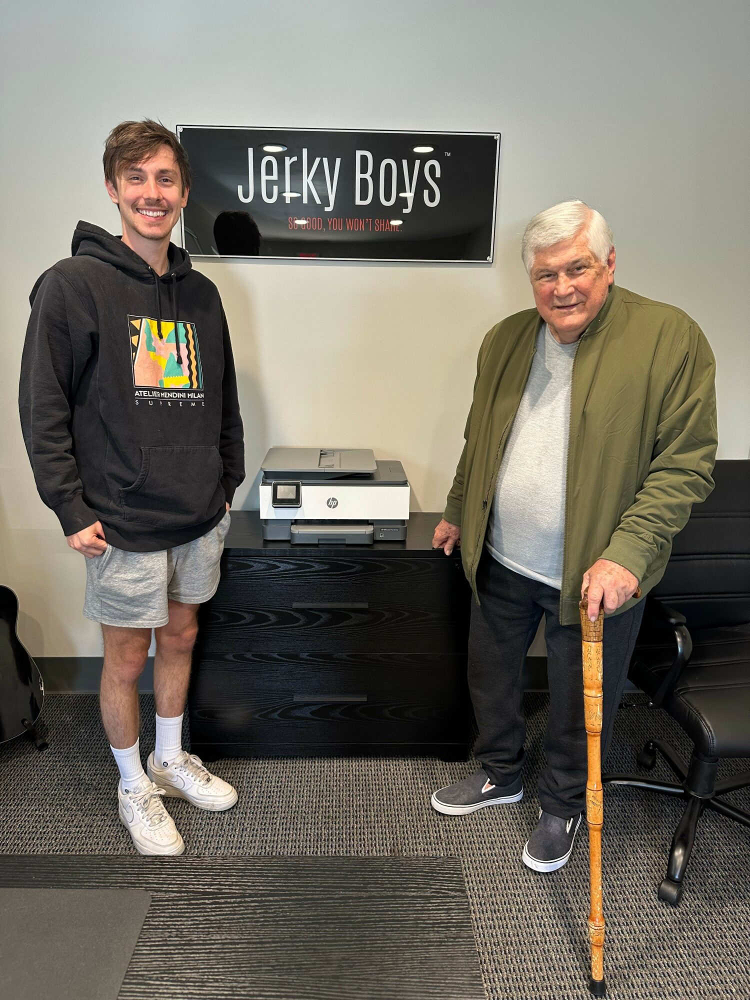 Two men standing in an office, one with a cane, in front of a sign that reads 'Jerkx Buys'.