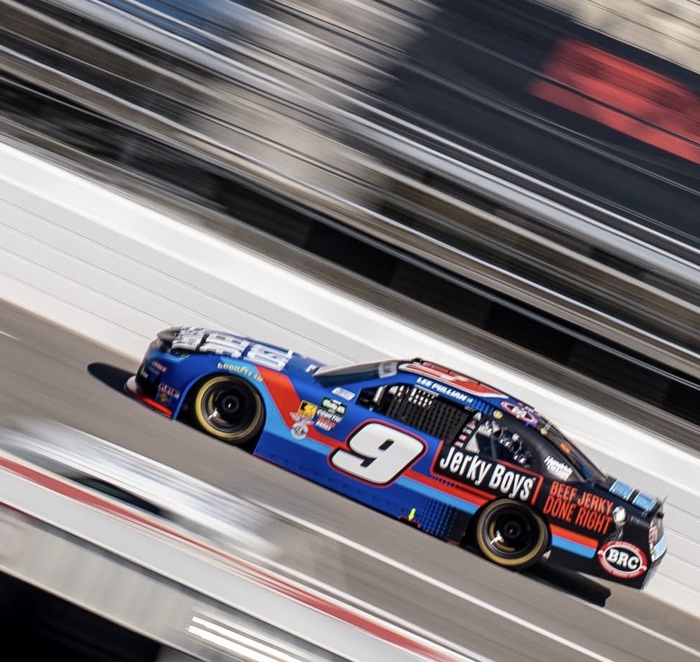 Race car with number 9 speeding on track, blurred background, blue and red livery, sponsor logos, black tires.
