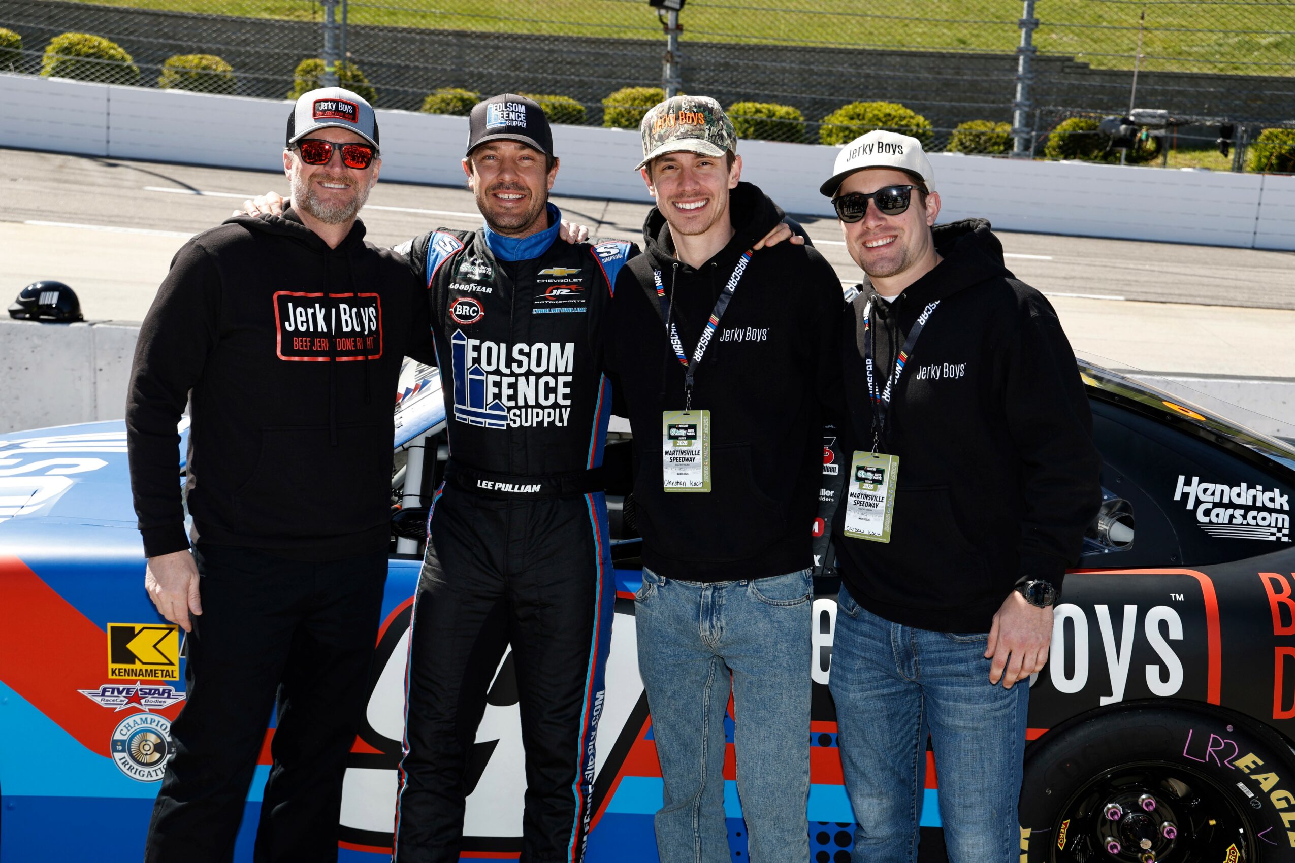 Four men standing together in front of a race car at a racing event, smiling and wearing casual clothing and caps.