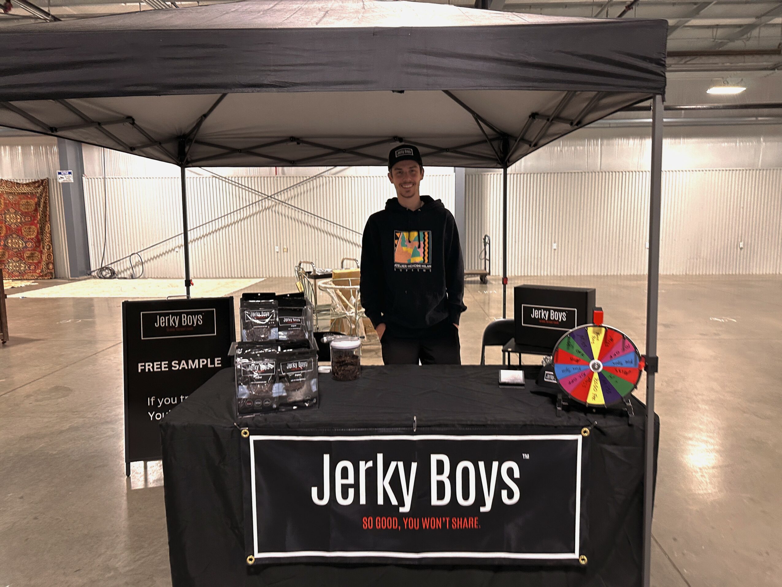 Person standing behind a booth with a Jerky Boys banner, a colorful spinning wheel, and various products inside a large indoor space.