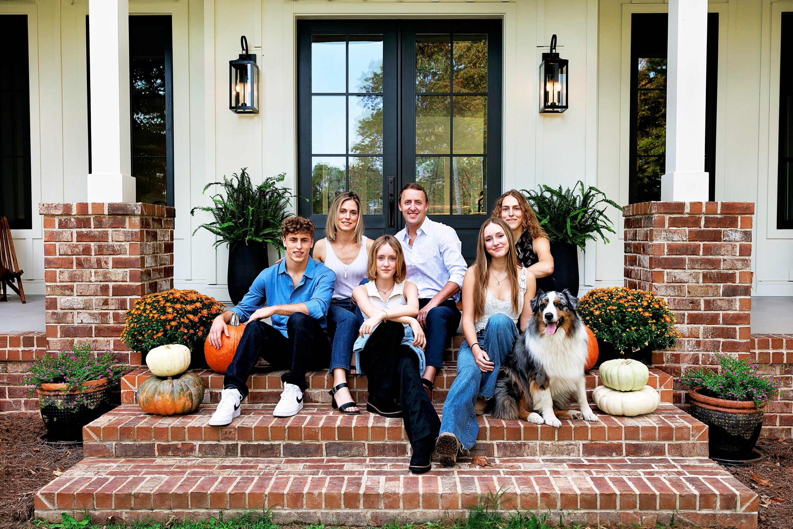 Family of six with a dog sitting on brick steps in front of a house with large window and potted plants, autumn decorations.