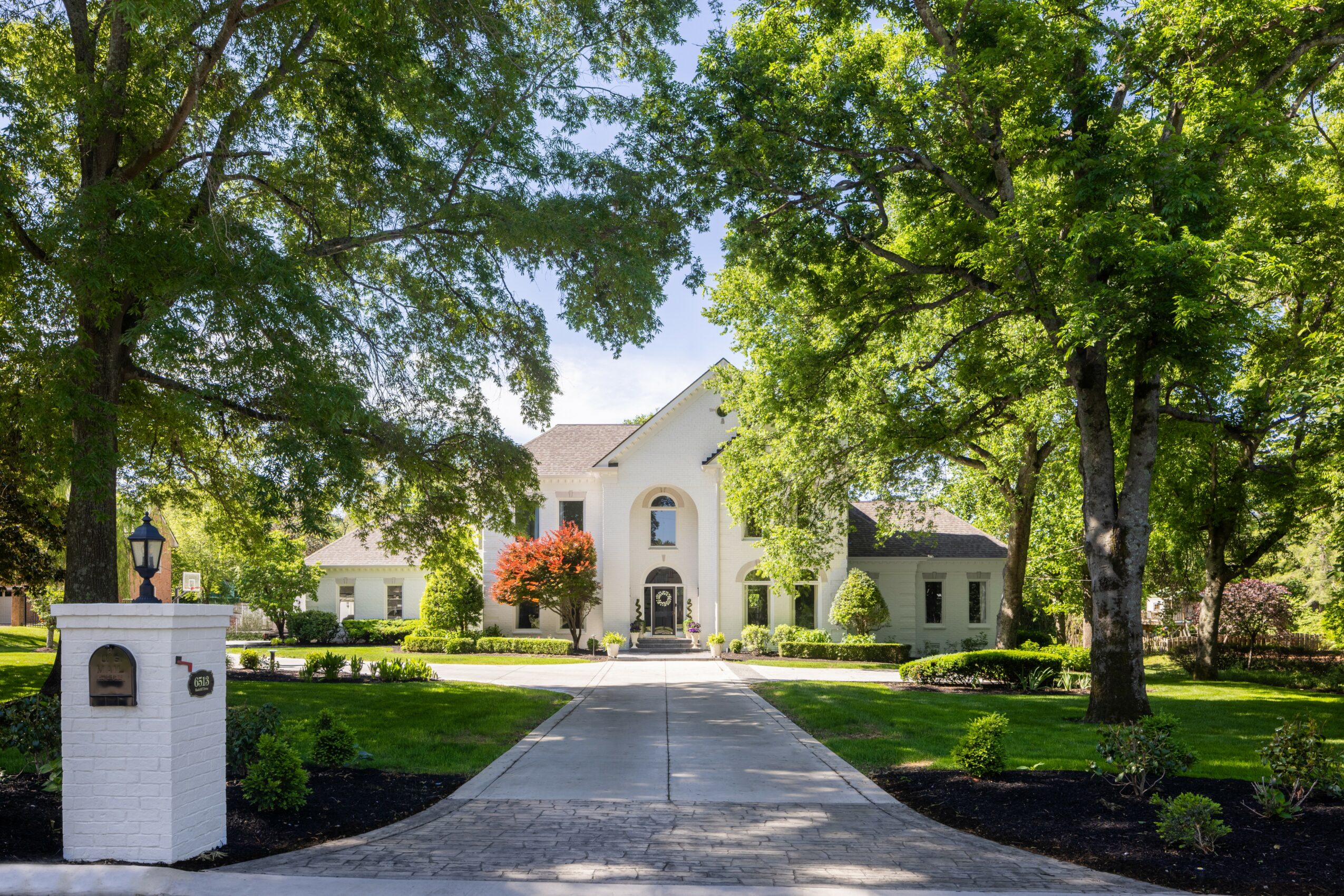 White house with arched entrance, surrounded by green trees and a well-maintained lawn, sunny day.