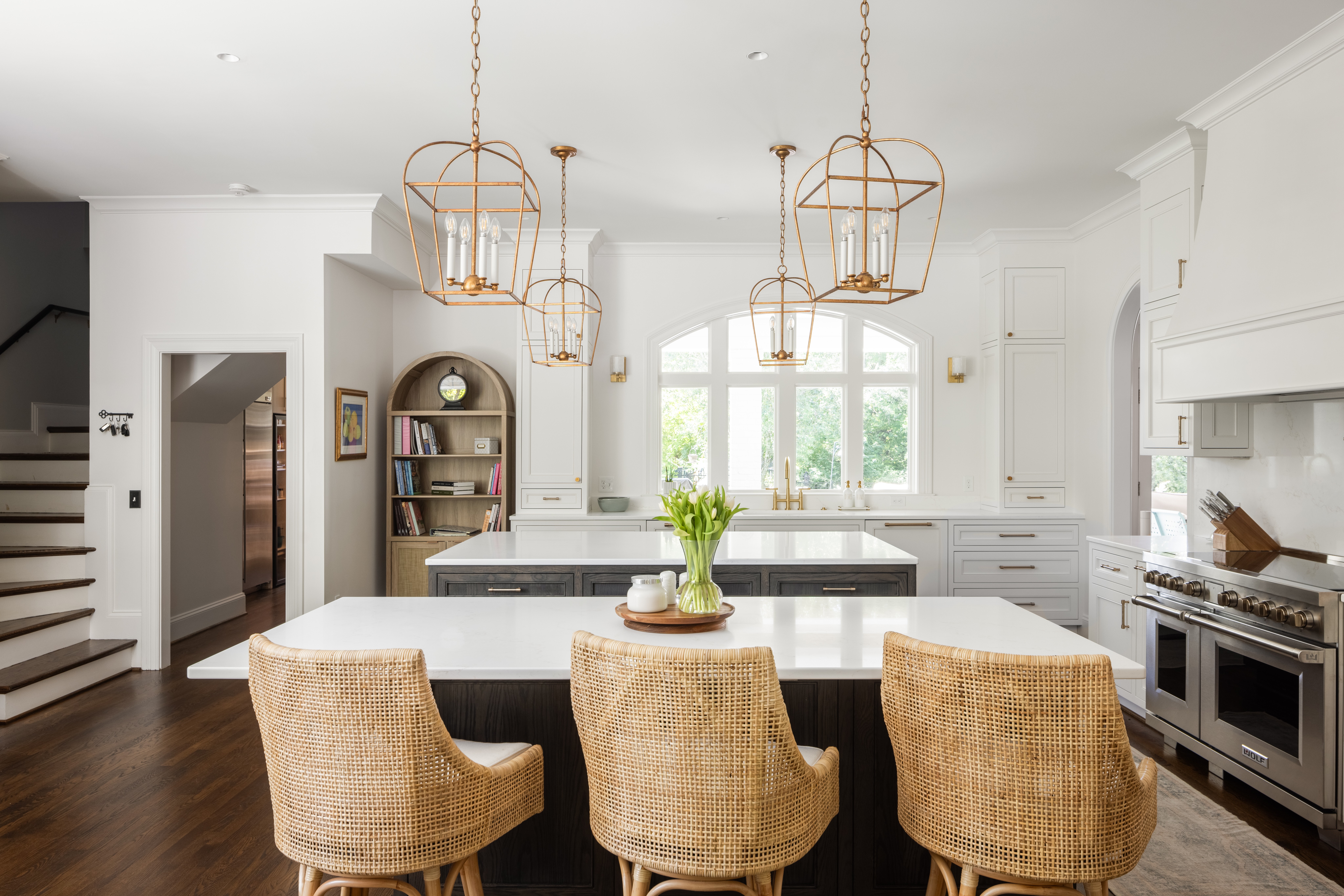 Bright kitchen with white cabinets, island, three beige chairs, hanging pendant lights, and a window with greenery outside.