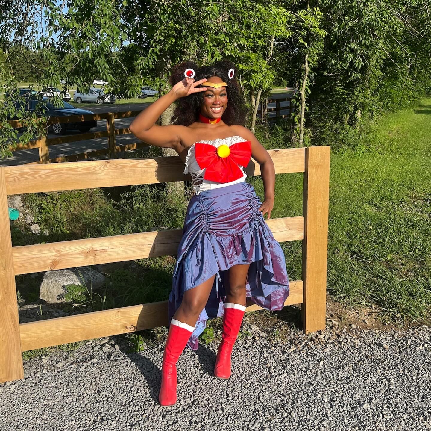Woman in festive costume with red, white, and blue colors, standing outdoors near a wooden fence, smiling, with trees and cars in background.