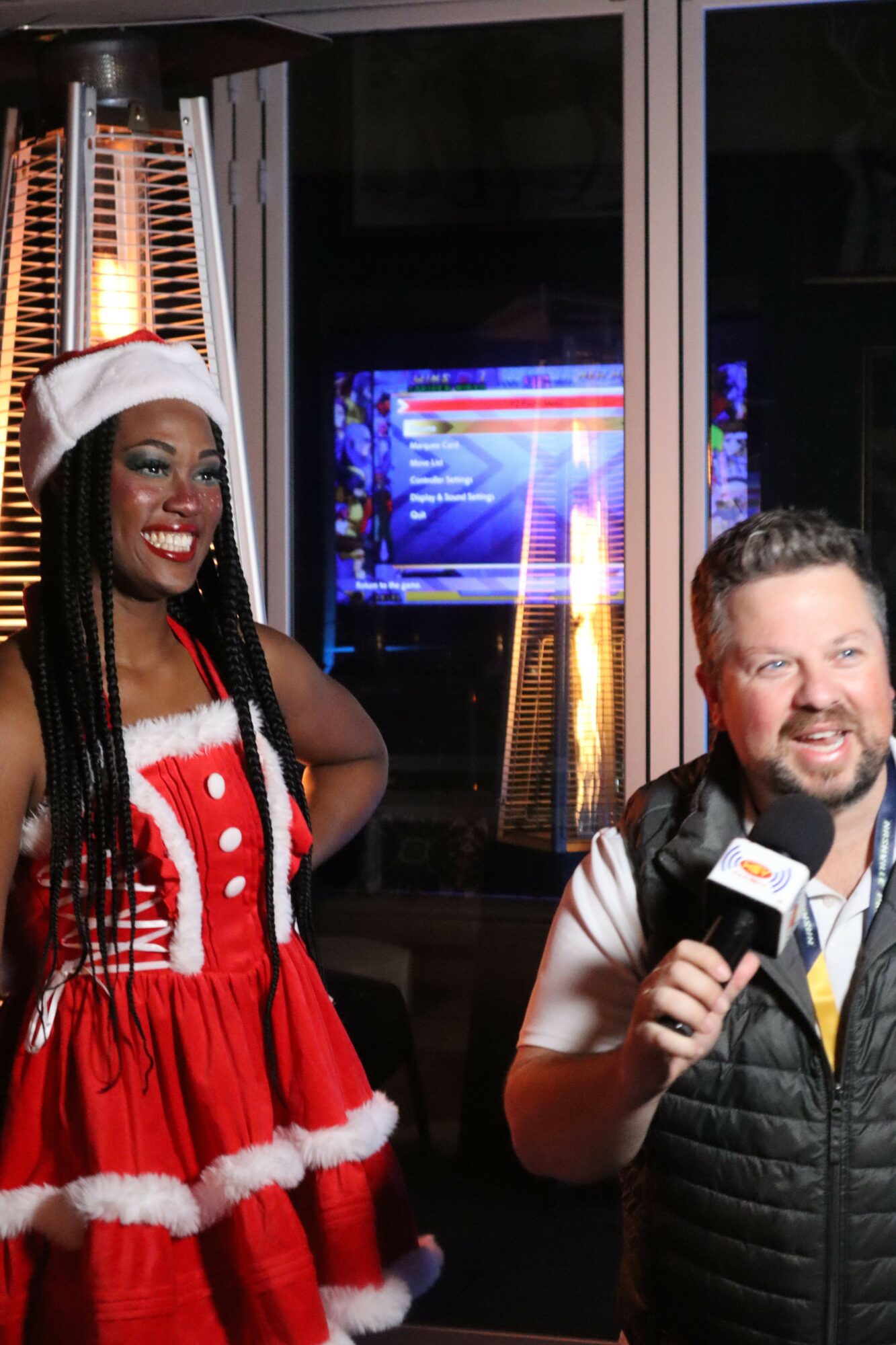 Two people smiling, one wearing a Santa hat and red dress, the other in a black jacket, in front of a window with colorful lights.
