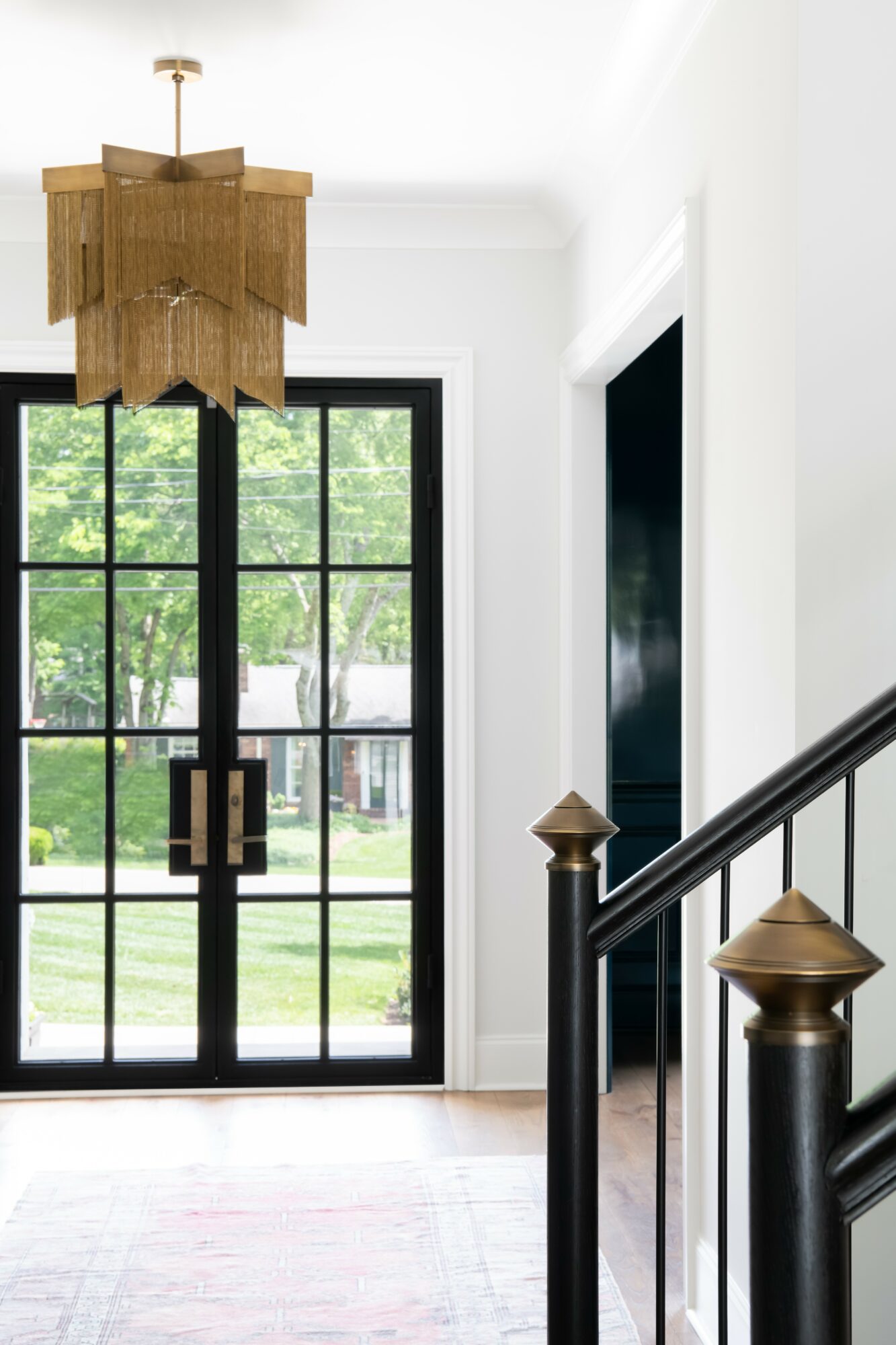 Entryway with black-framed glass door, white walls, staircase with black railing and gold accents, and a hanging light fixture.