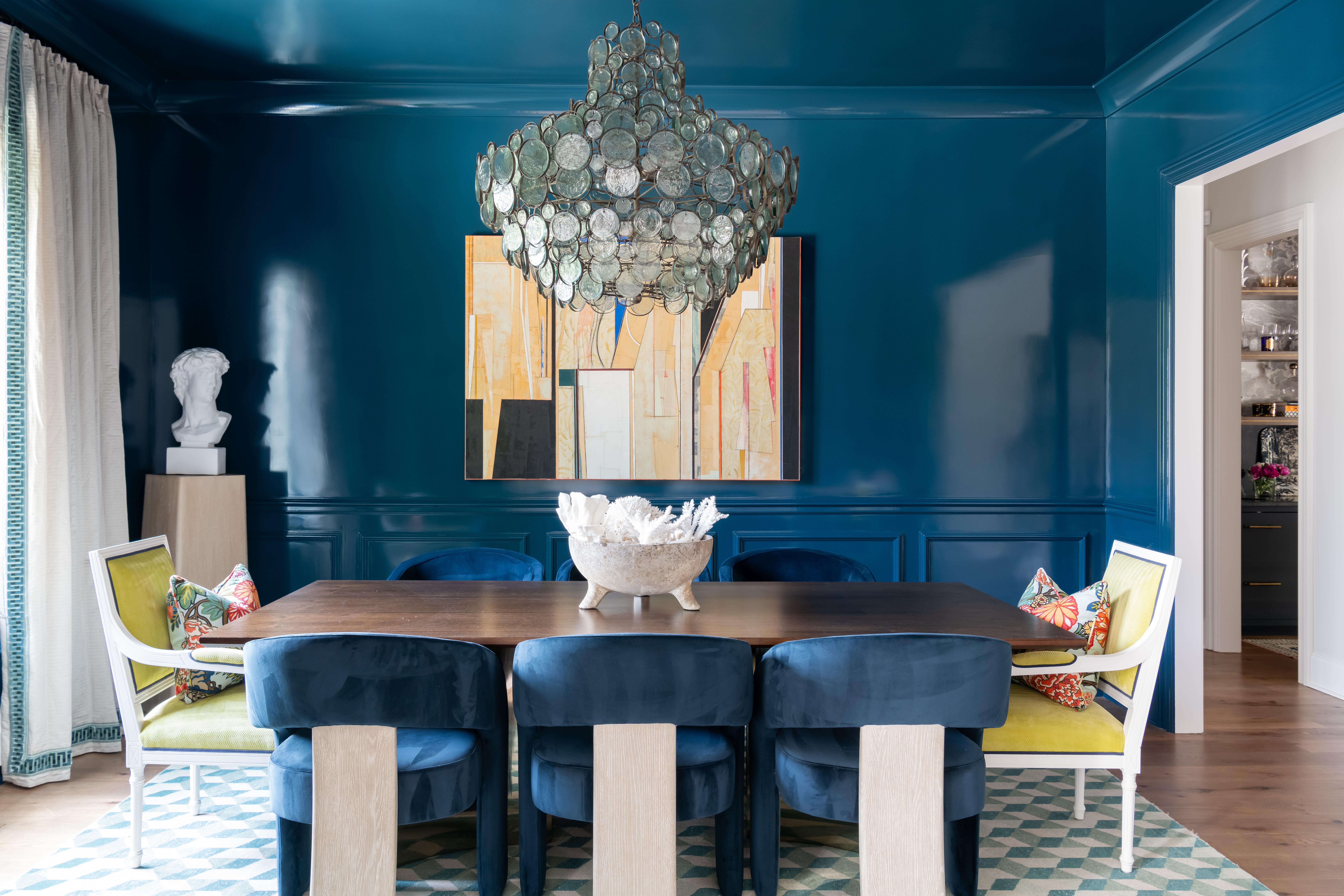 Dining room with blue walls, a chandelier, a round table with a decorative centerpiece, and colorful chairs.